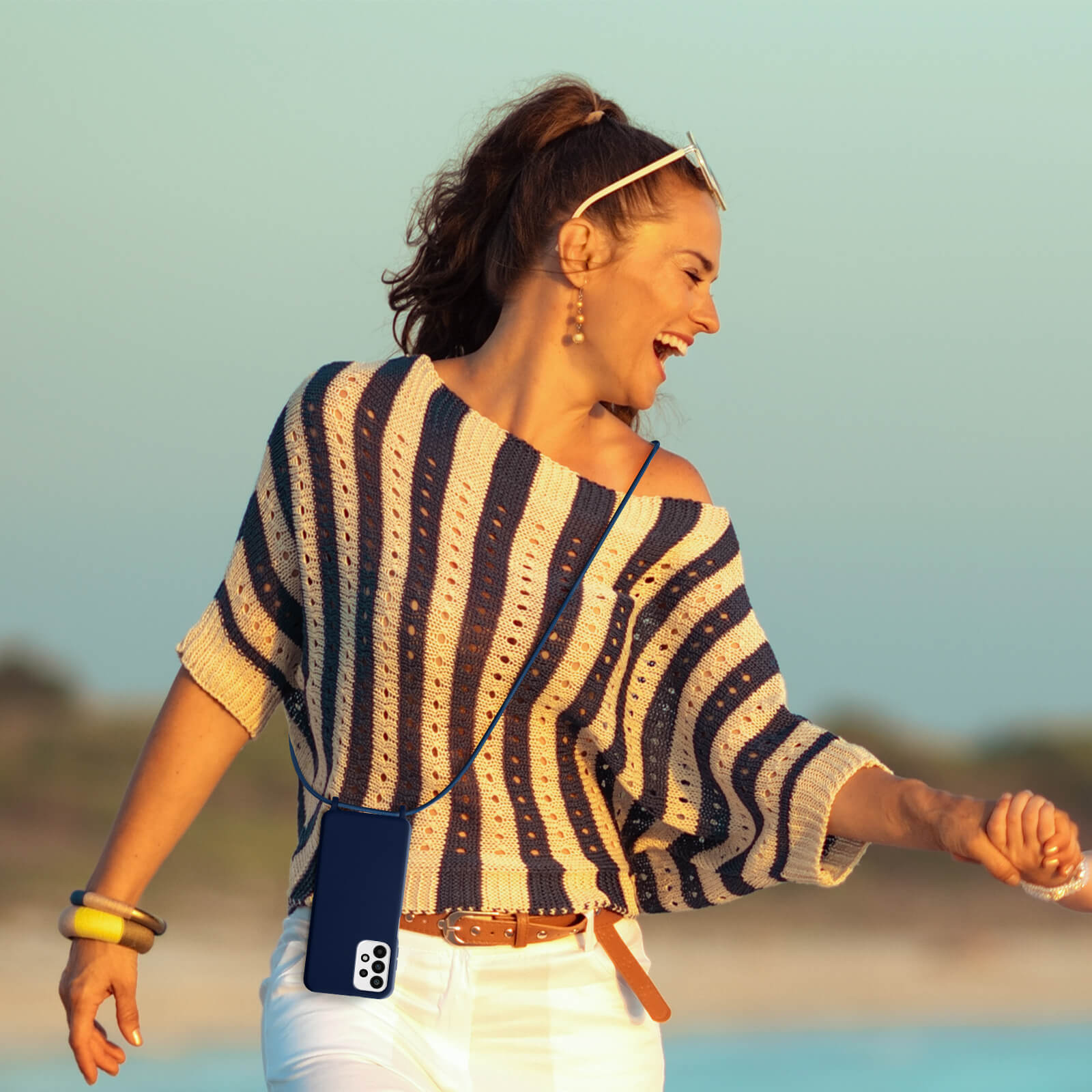 Femme portant une chemise rayée et un pantalon blanc, tenant un téléphone avec une coque. Flou.