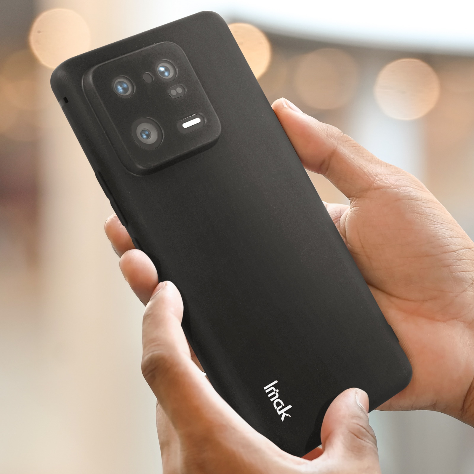 Una mano che tiene un telefono nero con un blocco fotocamera prominente. La custodia ha scritto 'imaki'.