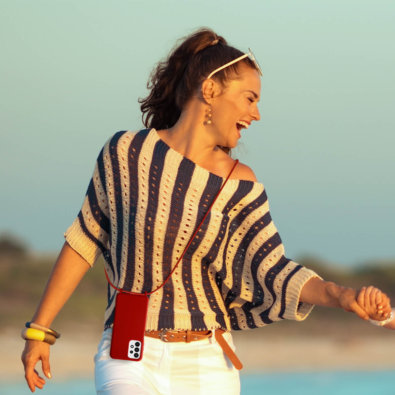 Vrouw in gestreepte shirt met telefoon aan riem, hand vasthoudend, lachend.
