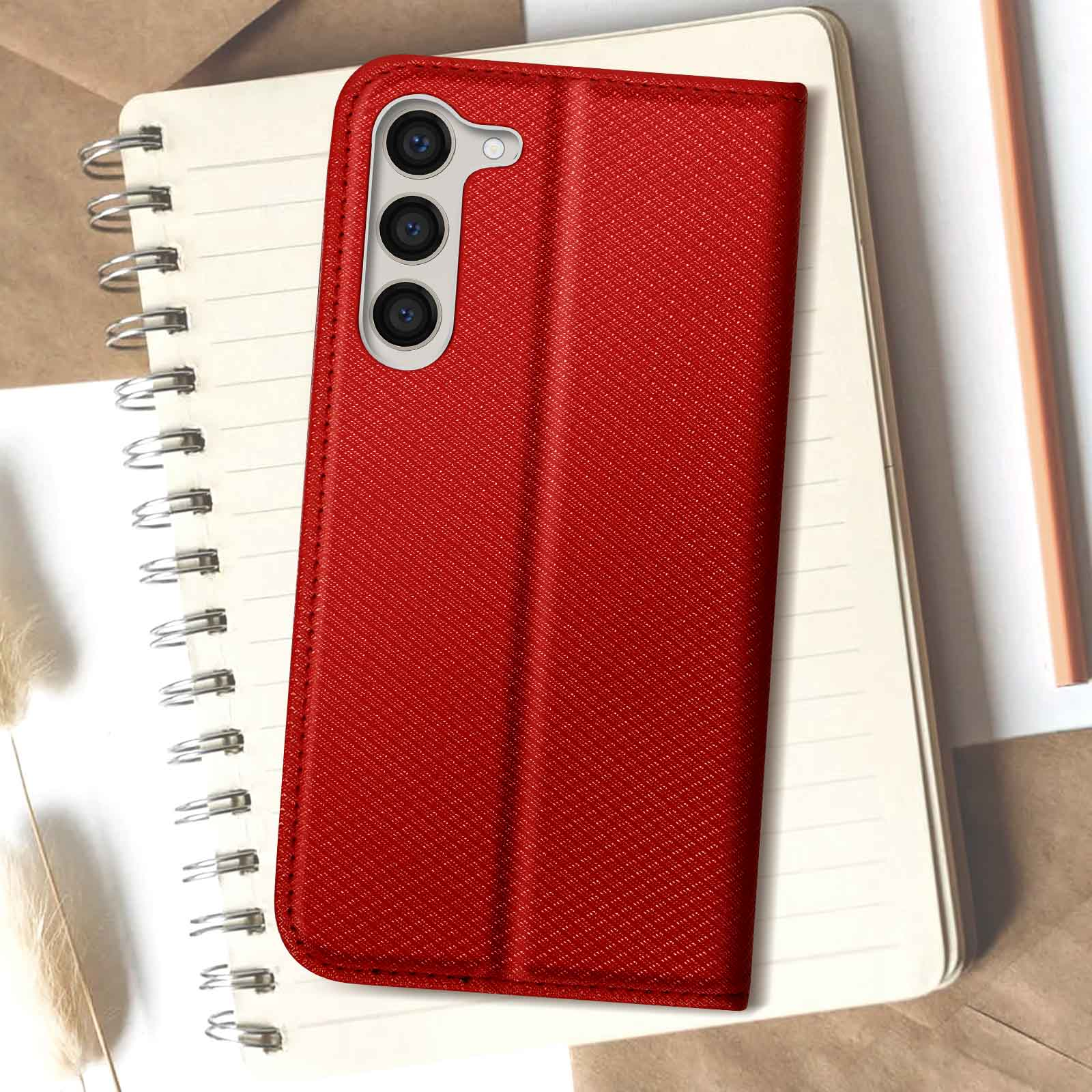 Eine rote Telefon-Geldbörsenhülle wird geöffnet auf einem Notizbuch gezeigt. Das Telefon hat drei Kameralinsen.