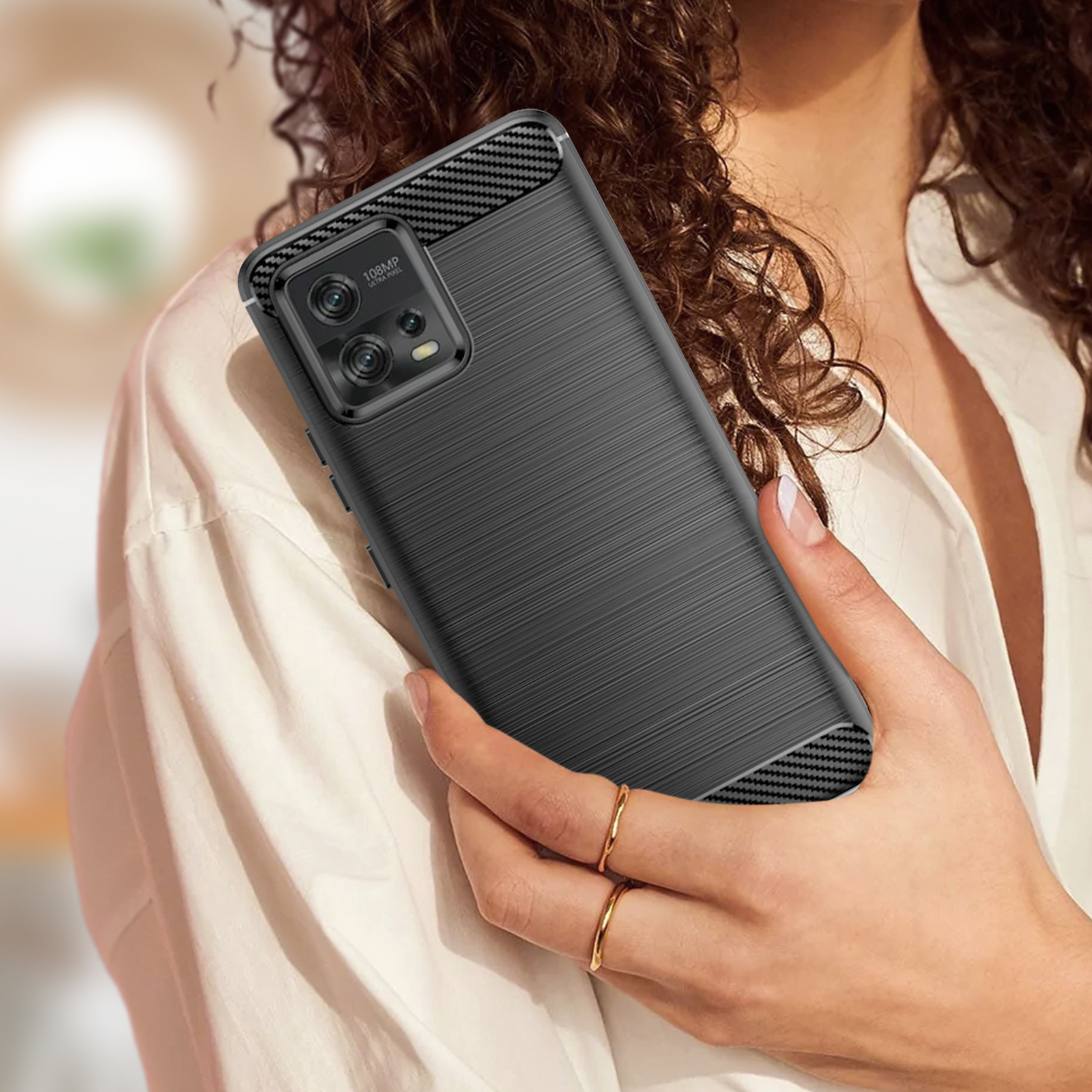Mani di donna che tengono un telefono nero con retro strutturato. Anelli d'oro, camicia bianca sullo sfondo.