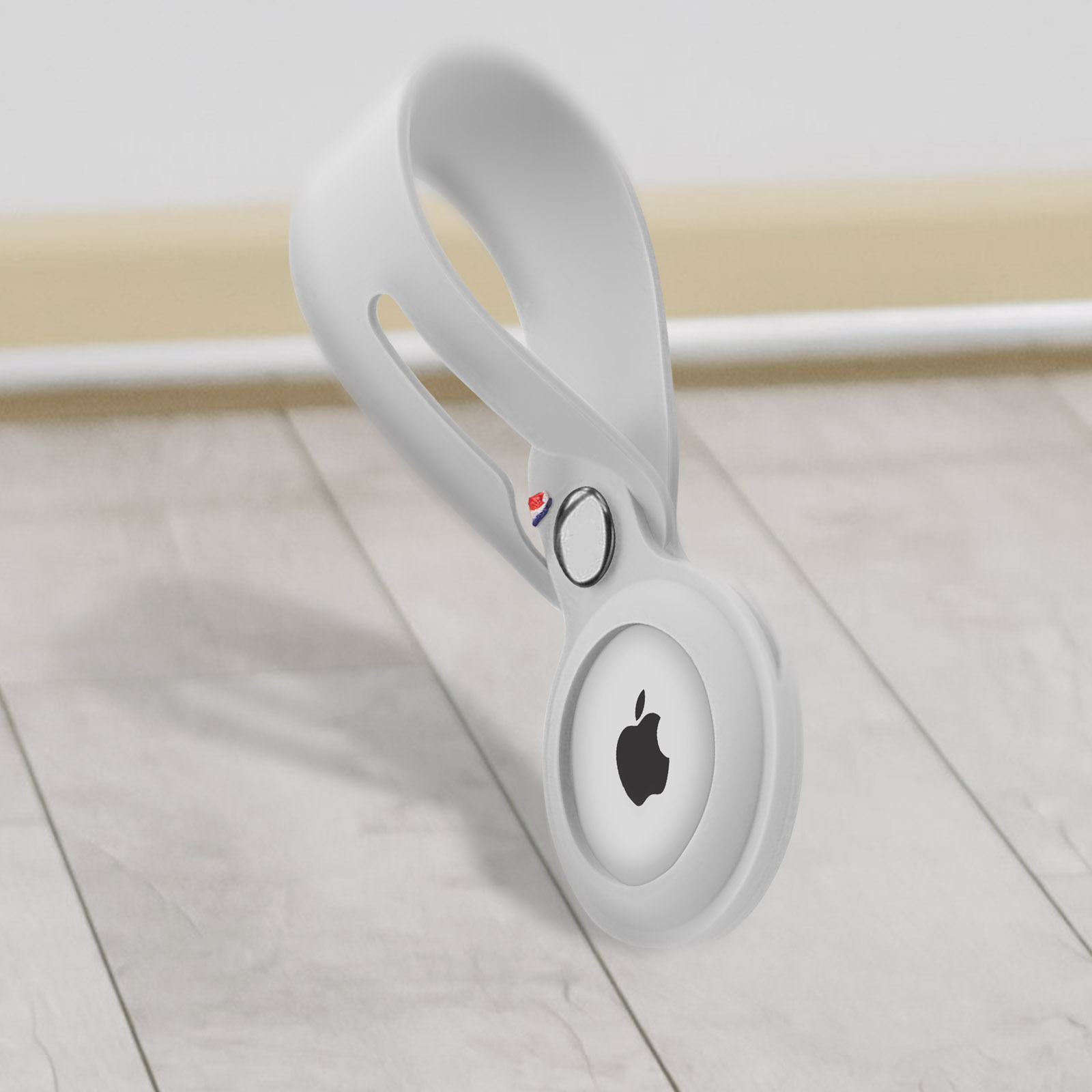 Un supporto bianco per Apple AirTag è mostrato, con il logo Apple. È su un pavimento in legno.