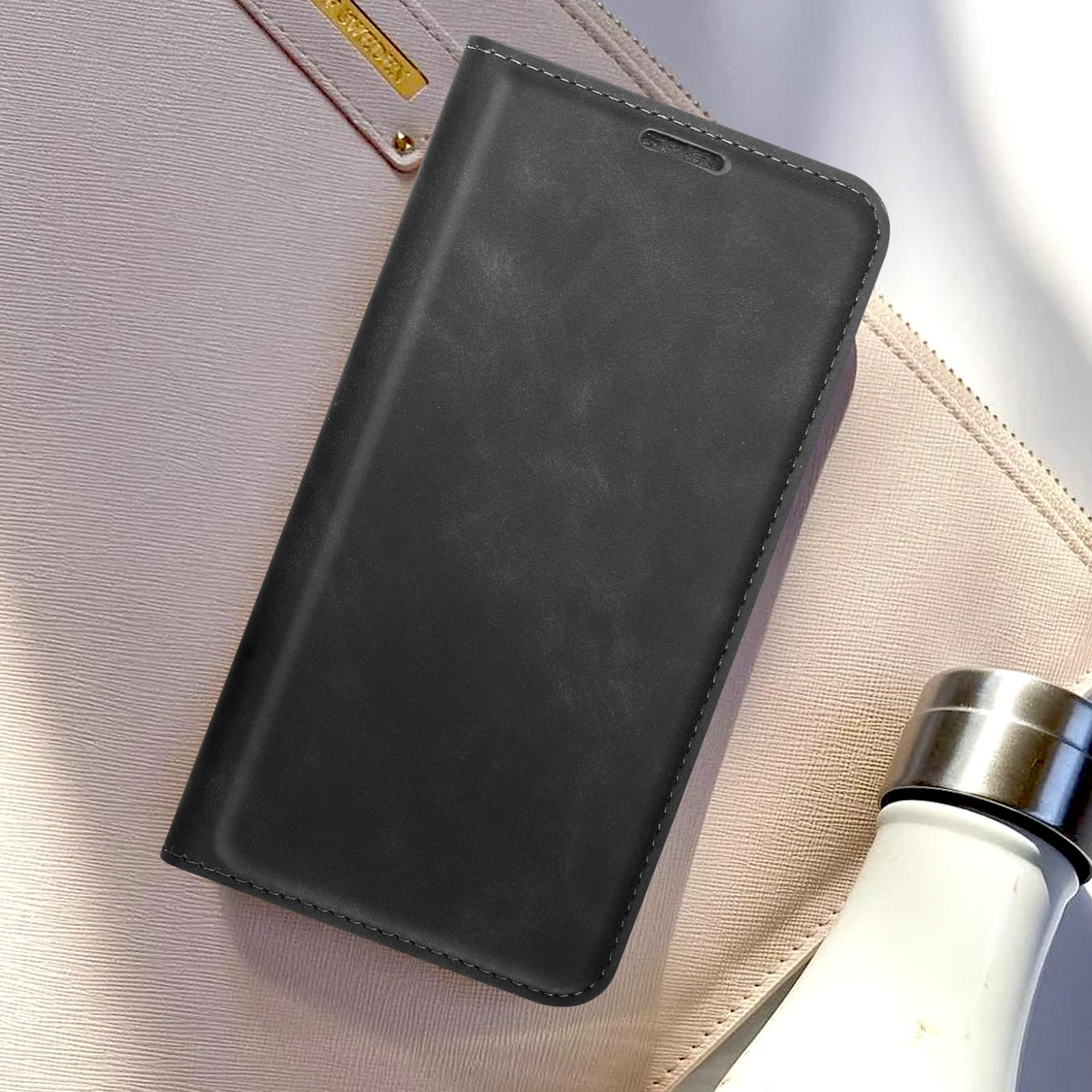 Schwarze Handyhülle auf einer beigen Handtasche. Eine weiße Flasche im Hintergrund.