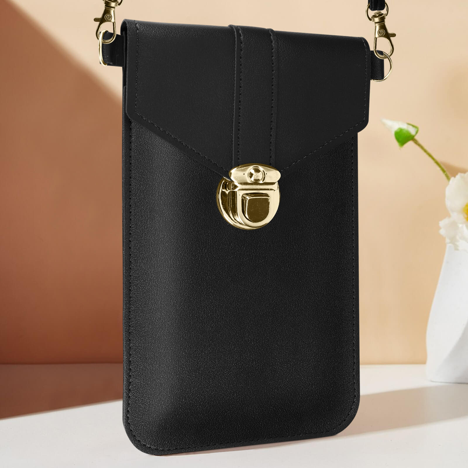 Borsa per telefono nera con chiusura e catena dorate. Superficie bianca, sfondo beige con fiore.