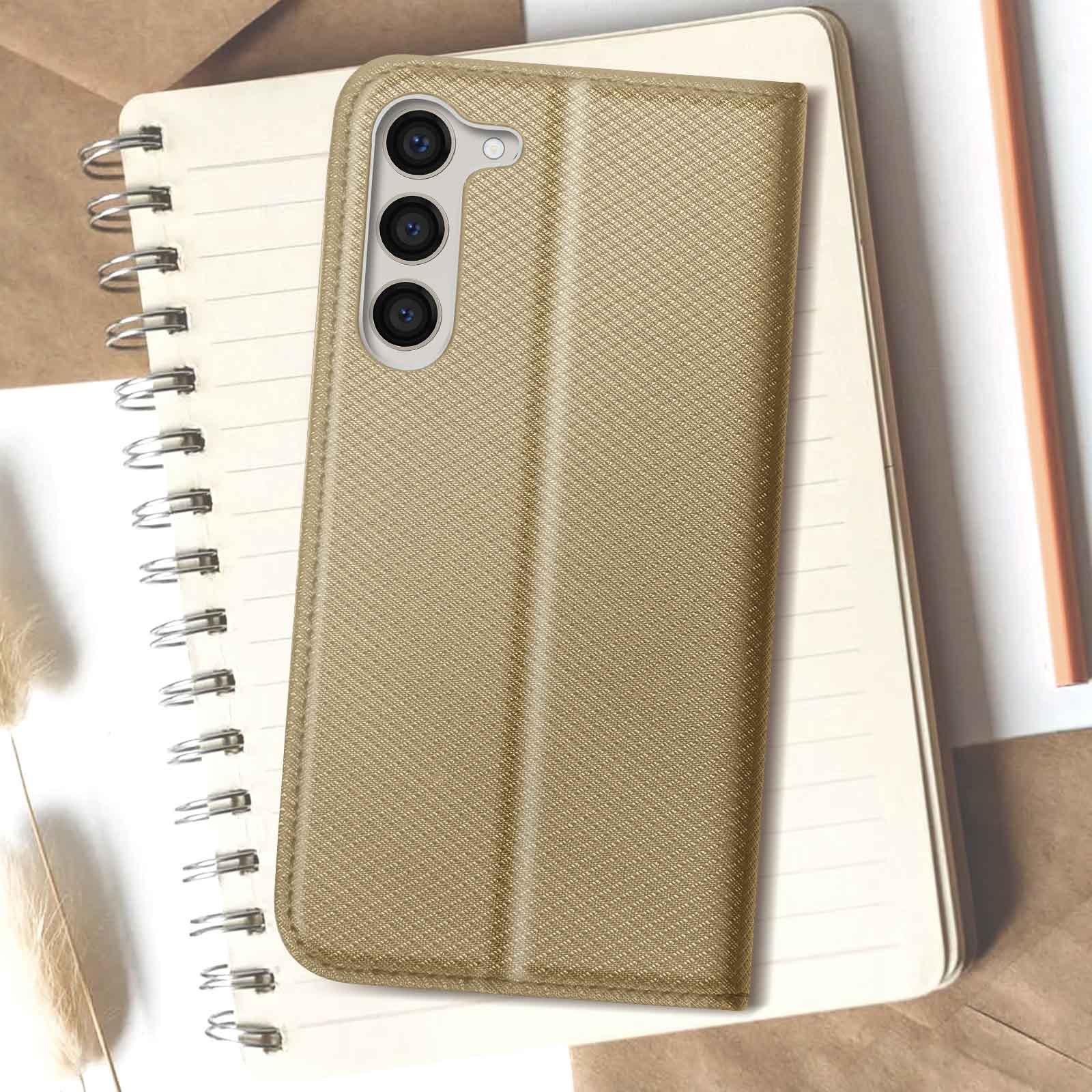 Eine goldfarbene Handyhülle auf einem Notizbuch mit einem Stift. Die Hülle ist geschlossen und strukturiert.