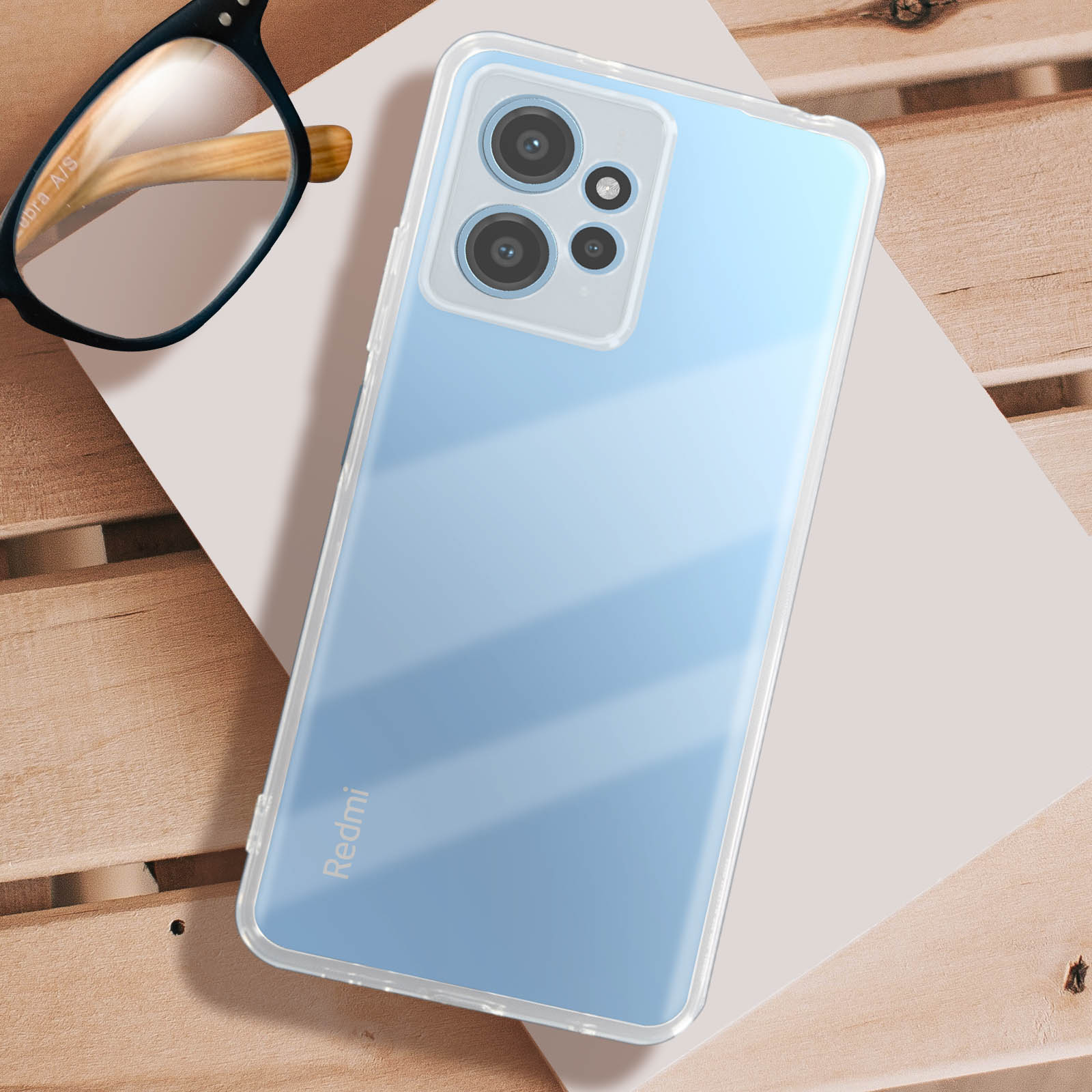 Un telefono in una custodia trasparente, con il logo Redmi. Un paio di occhiali accanto su una superficie di legno.