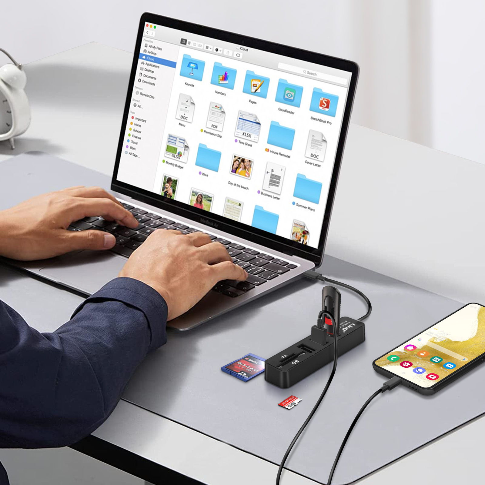 Una persona che lavora su un laptop con un hub USB, telefono e schede SD collegati. La scena è al chiuso.