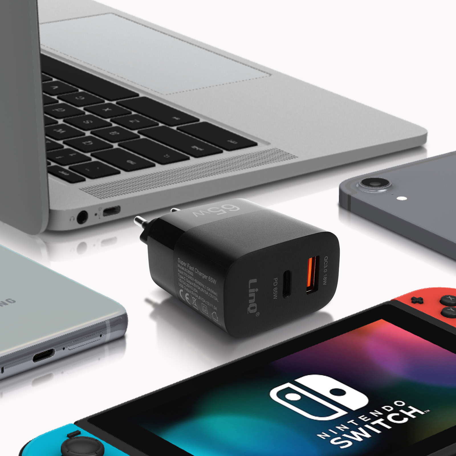 Un cargador negro está sobre una mesa. Junto al cargador hay varios aparatos electrónicos como un portátil y una Nintendo Switch.