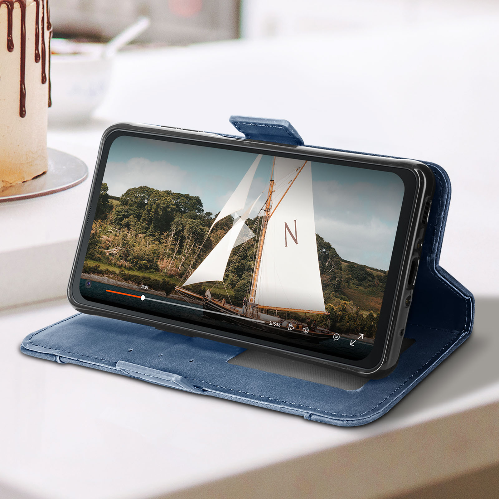 Un telefono riproduce un video su una custodia blu. C'è una torta nell'angolo su una superficie bianca.