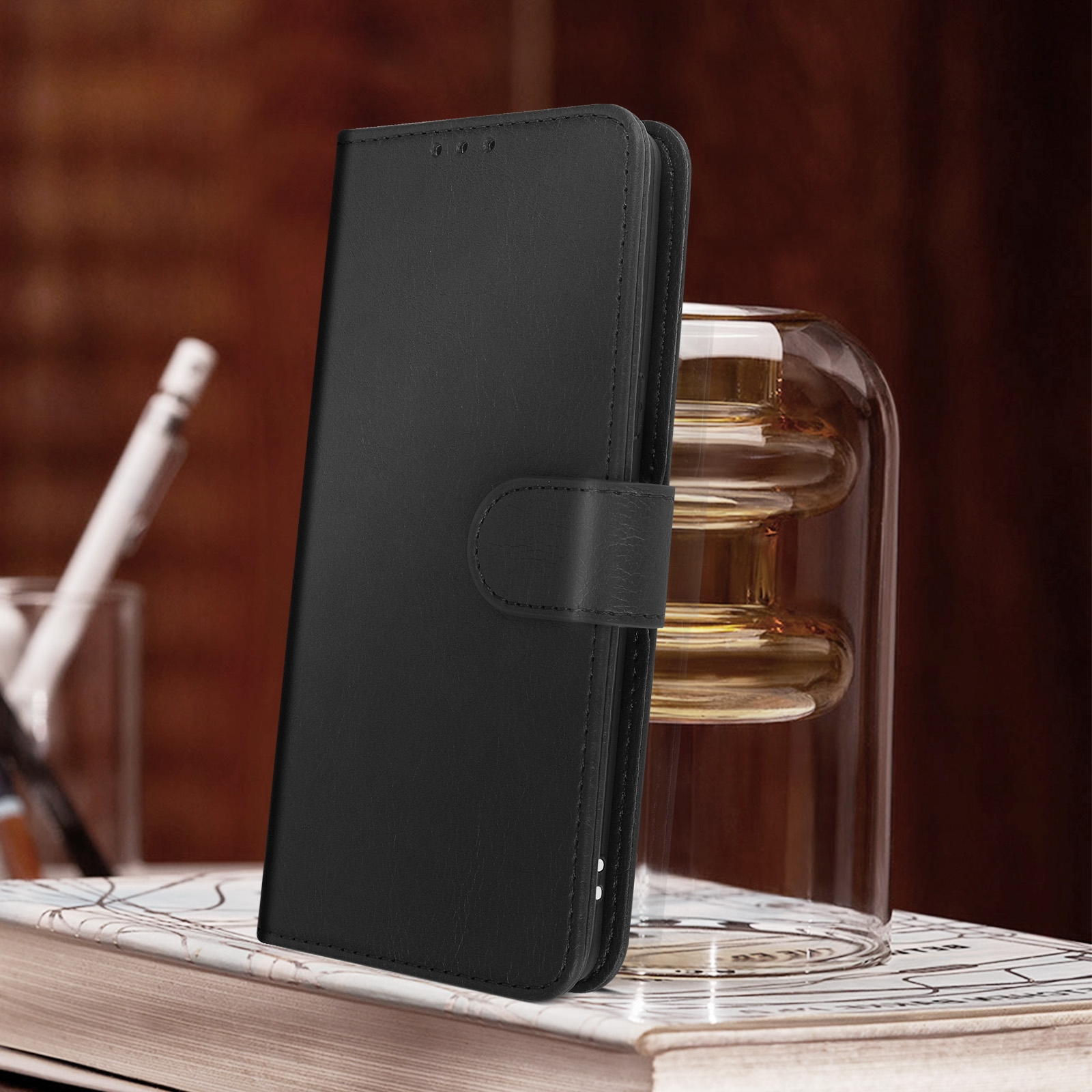 Custodia nera per telefono su un libro. Un vaso di vetro e una matita sfocati sullo sfondo.