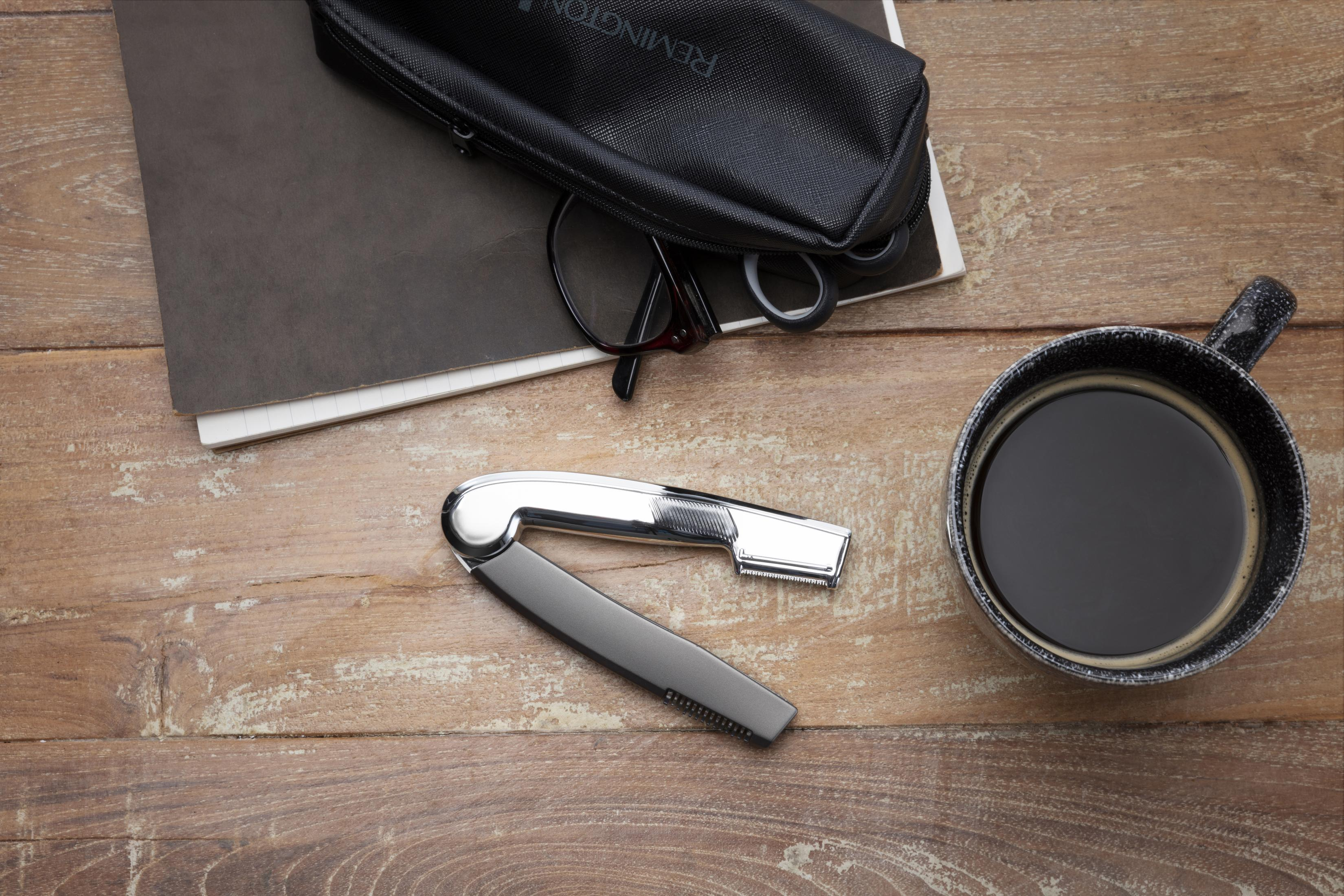 Vista dall'alto di strumenti per la cura della persona su una superficie di legno: rasoio, taccuino, occhiali e tazza di caffè.