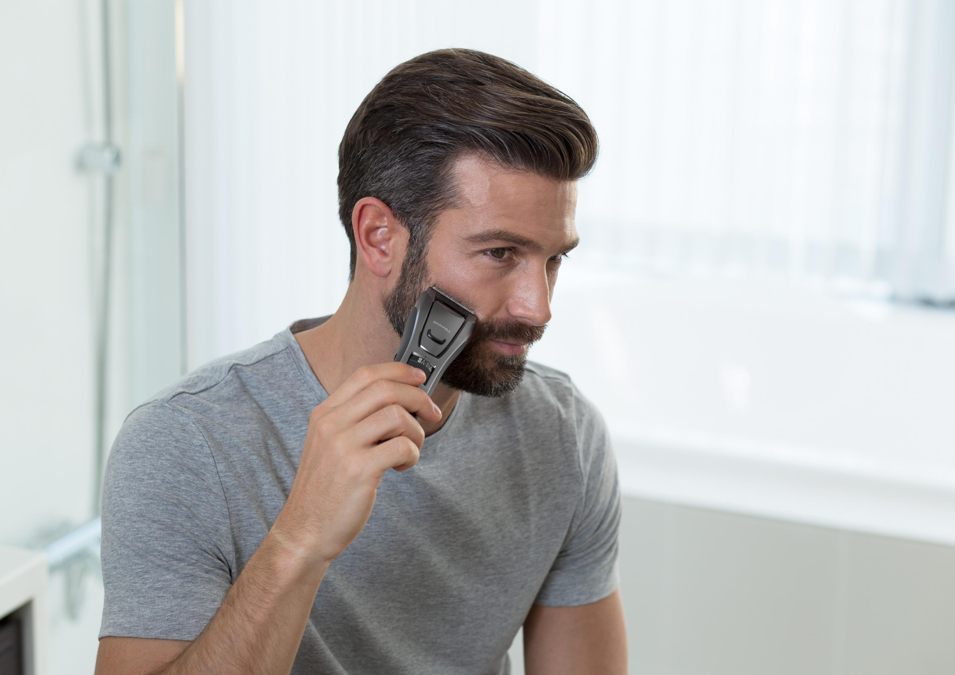 Mężczyzna goli brodę golarką elektryczną. Jest w łazience z lustrem.