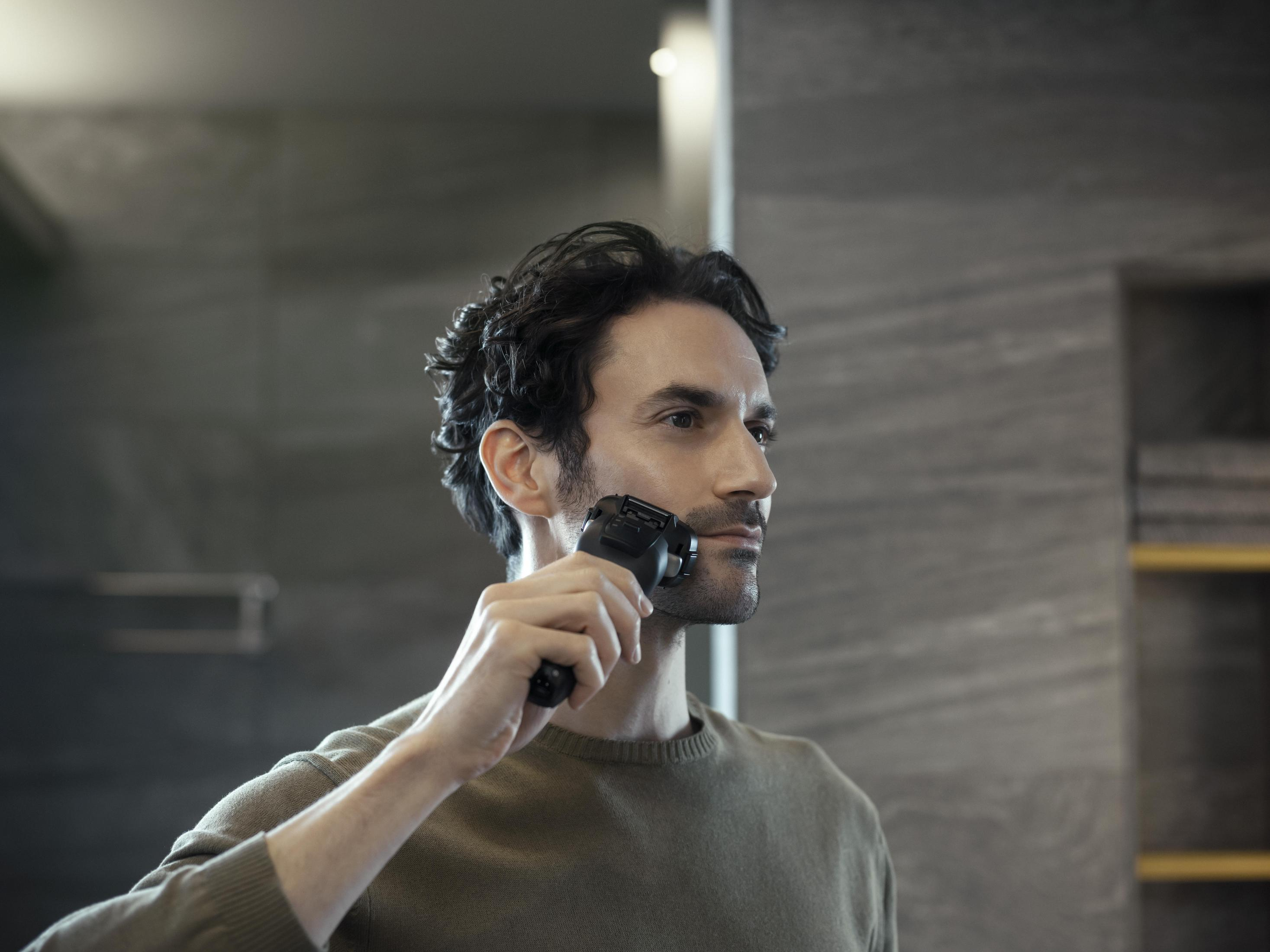 Mężczyzna goli twarz w lustrze. Używa golarki elektrycznej w łazience.