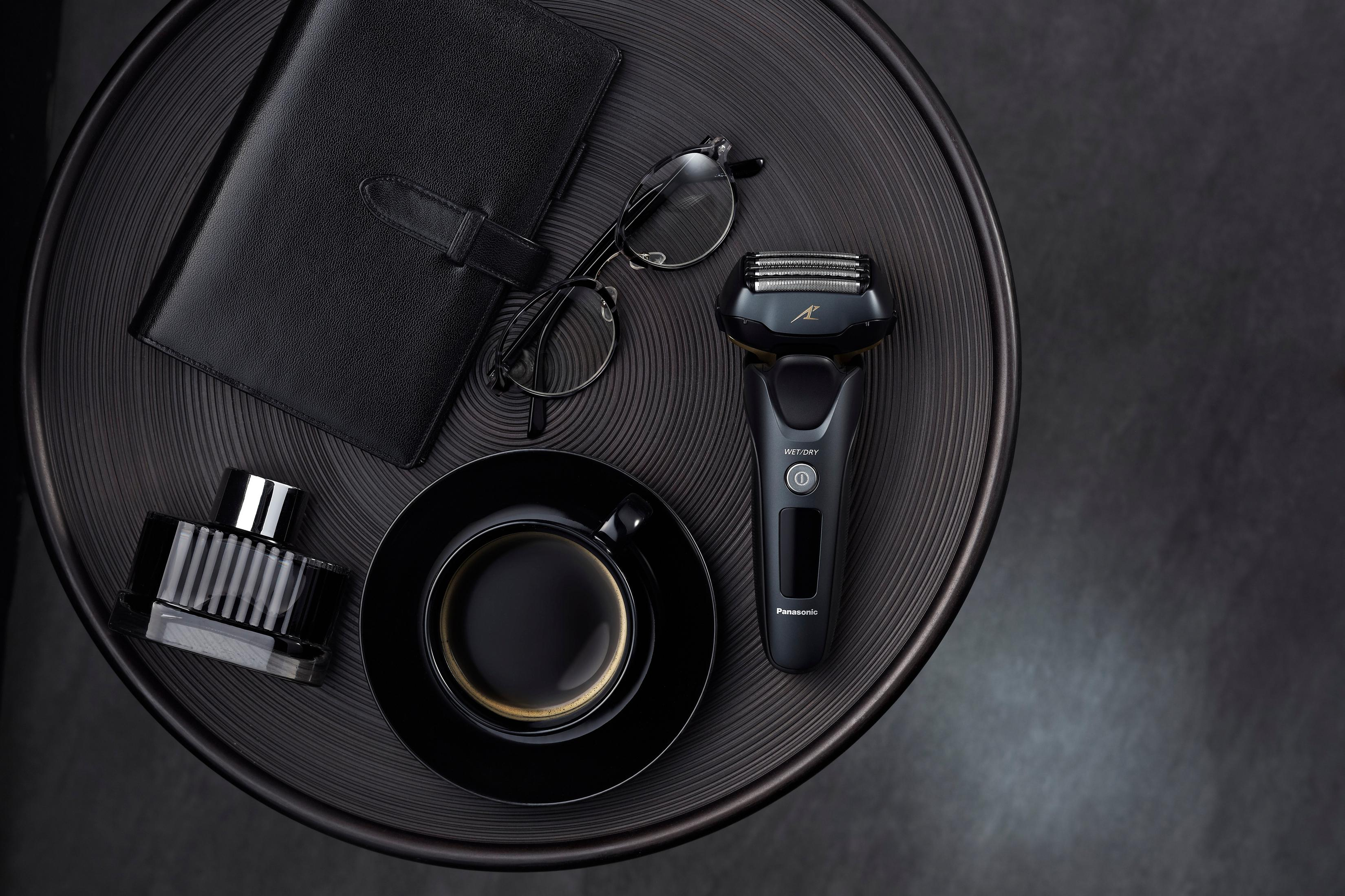 Schwarze Pflegeaccessoires auf einem schwarzen Tisch. Enthält einen Rasierer, eine Brille, ein Notizbuch, Kaffee und Parfüm.
