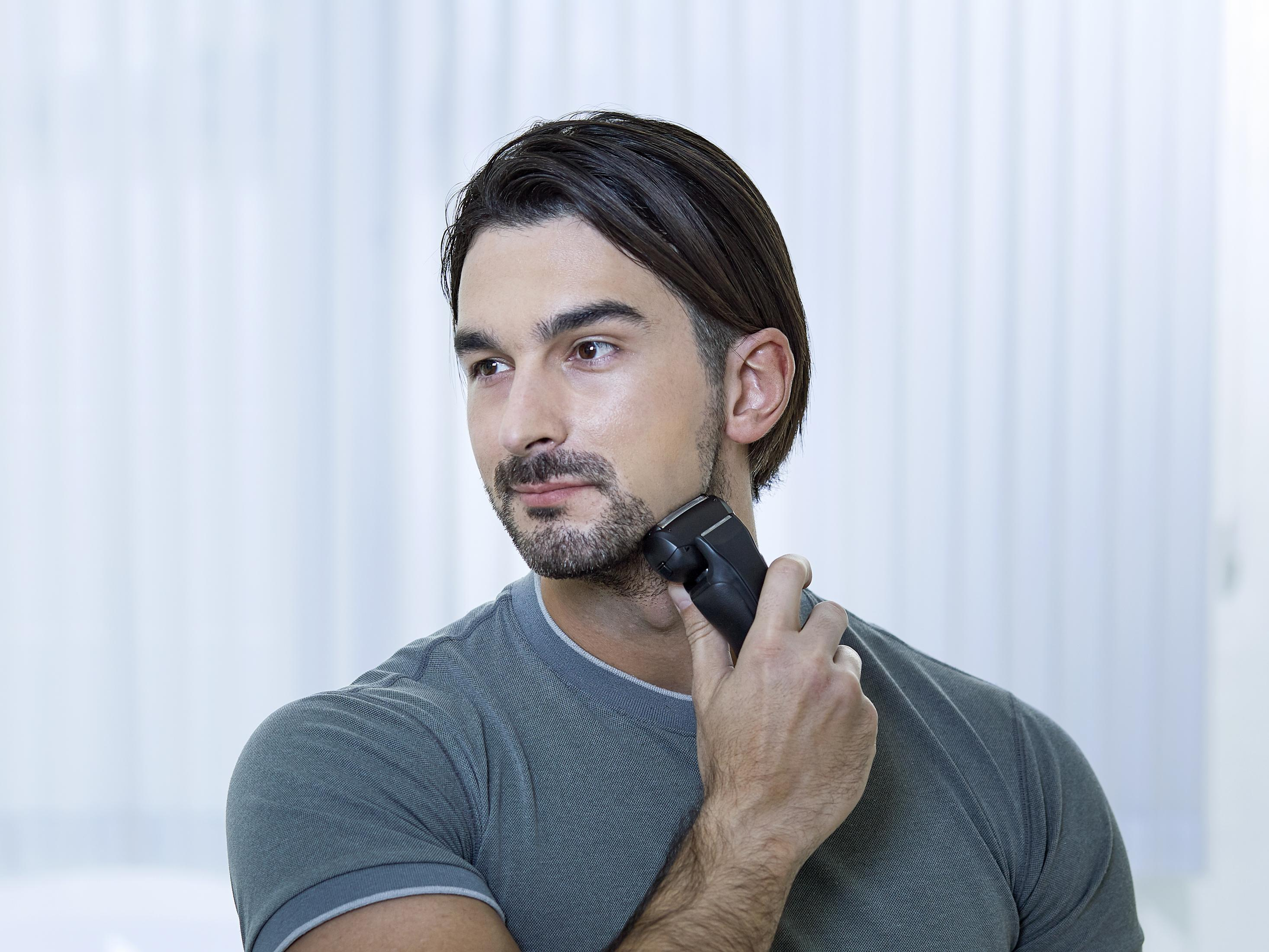 Mężczyzna goli brodę trymerem. Ma na sobie szarą koszulkę, patrzy w bok. Tło to biała ściana.