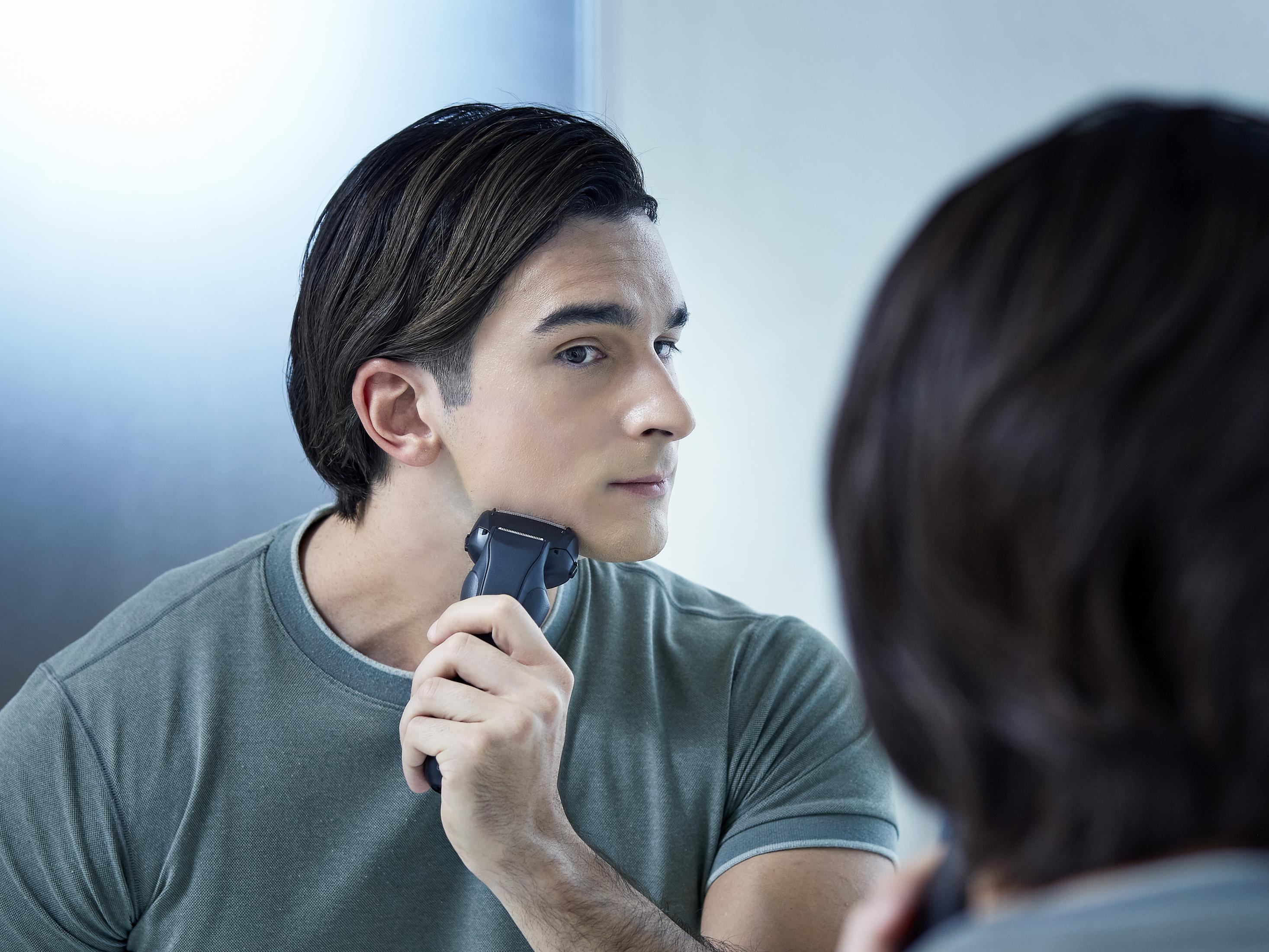 Mężczyzna goli twarz golarką elektryczną, patrząc w lustro. Tłem jest łazienka.