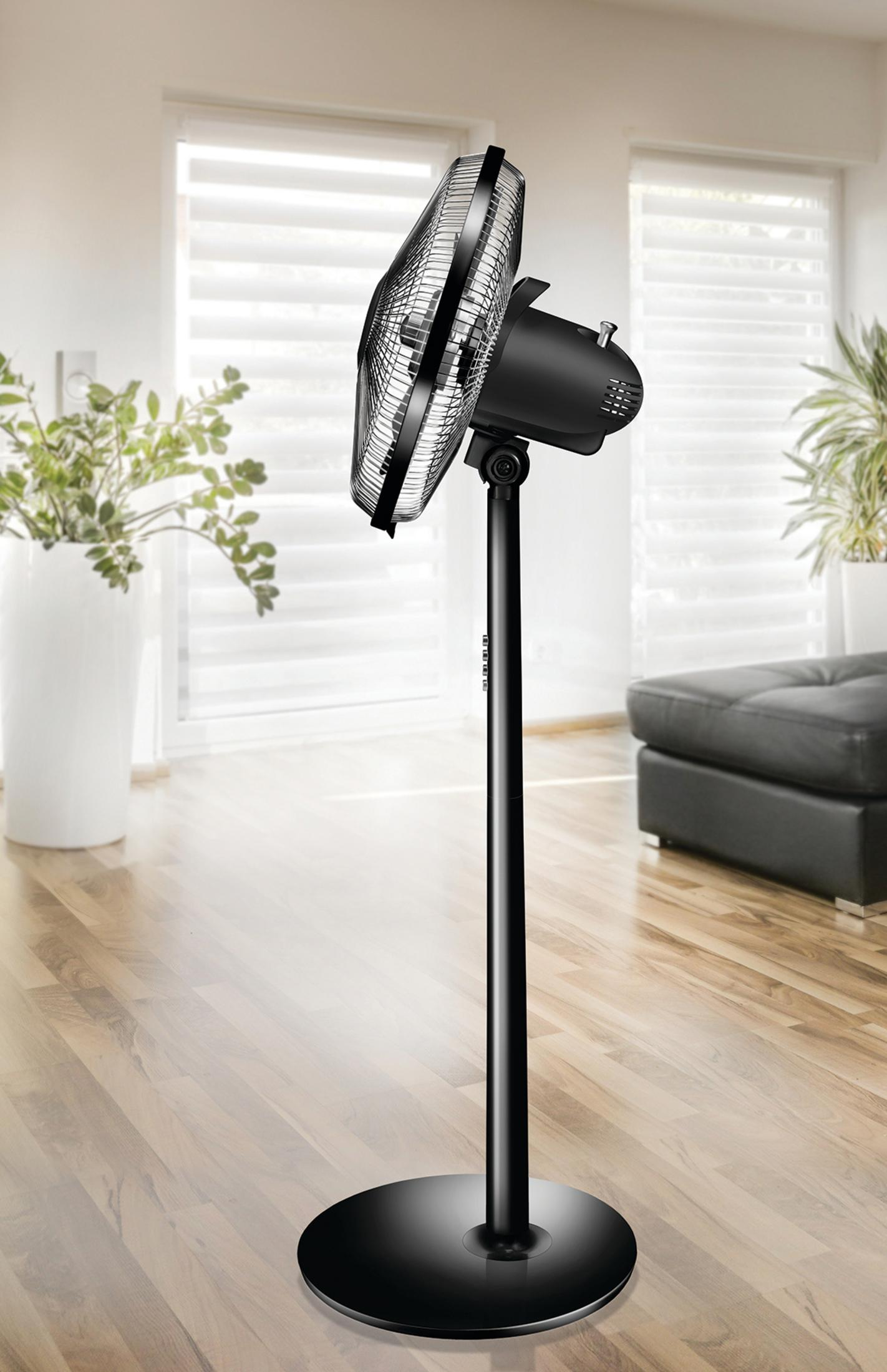 Czarny wentylator stojący na drewnianej podłodze. W tle duże okna.
