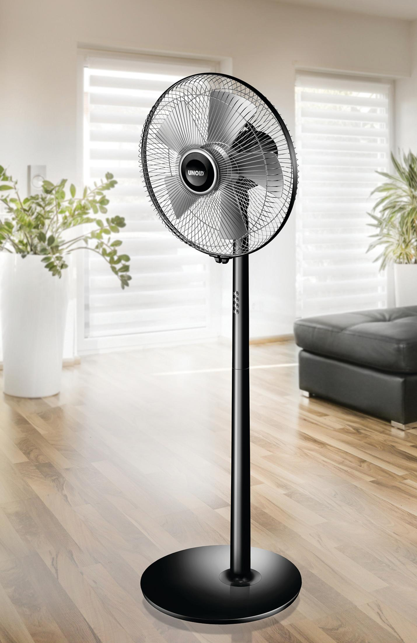 Czarny wentylator stojący w jasnym pokoju. Stoi na drewnianej podłodze, z białymi ścianami i czarną kanapą.