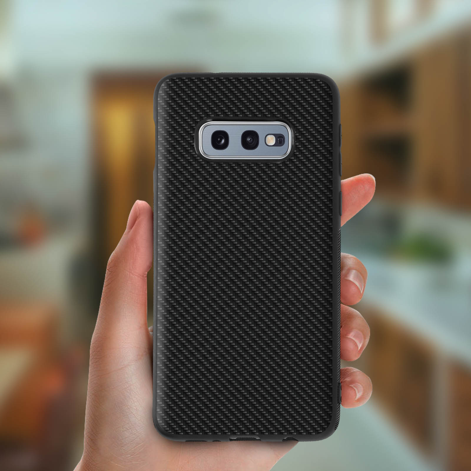 Una mano tiene un telefono con una custodia in stile fibra di carbonio. Lo sfondo è una cucina sfocata.