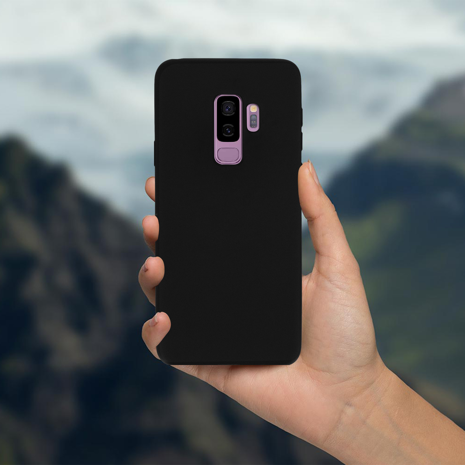 Hand hält schwarzes Telefon, schwarze Hülle. Hintergrund verschwommen, Berge.