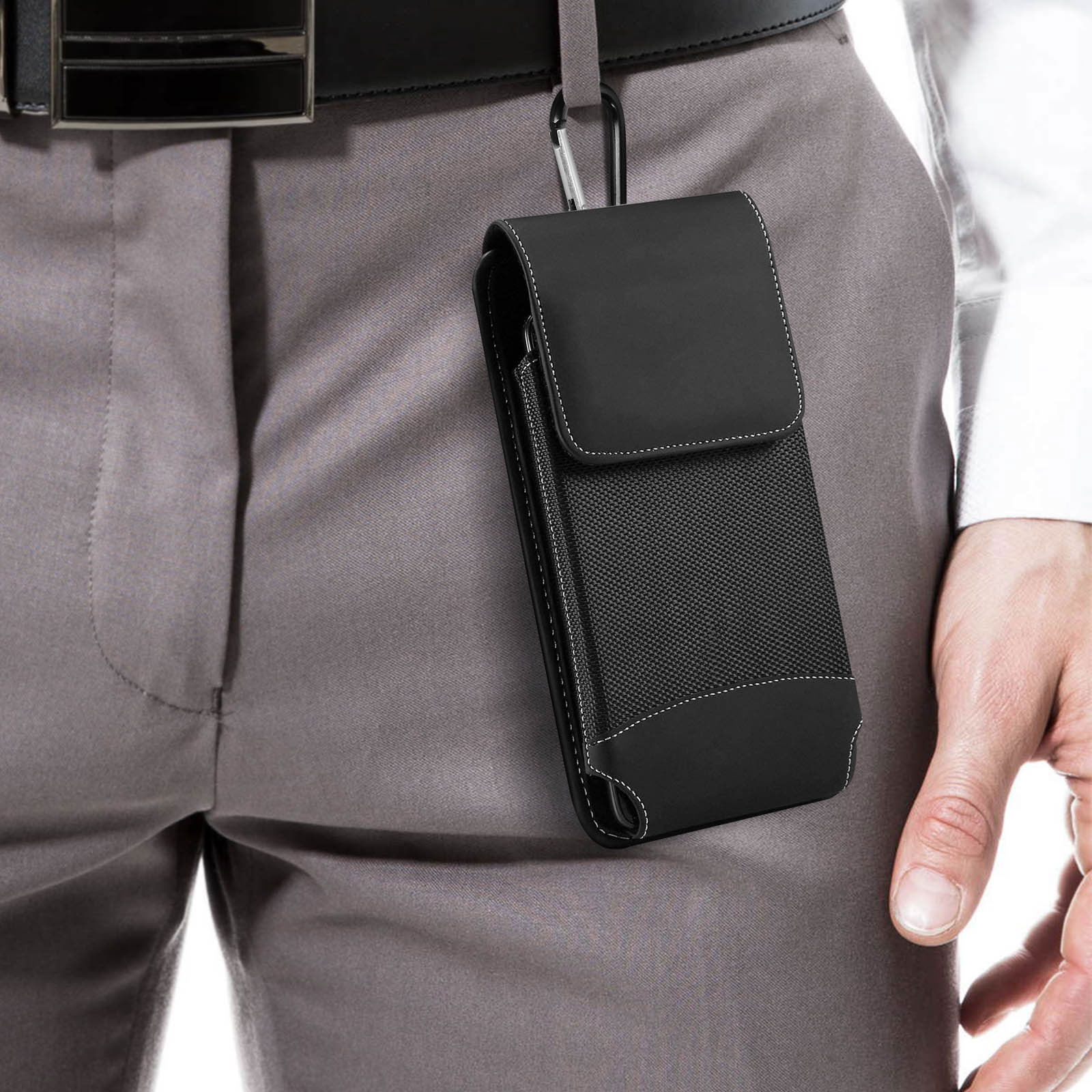 Custodia nera per telefono attaccata al passante della cintura, tenuta da una mano, sfondo pantaloni grigi.