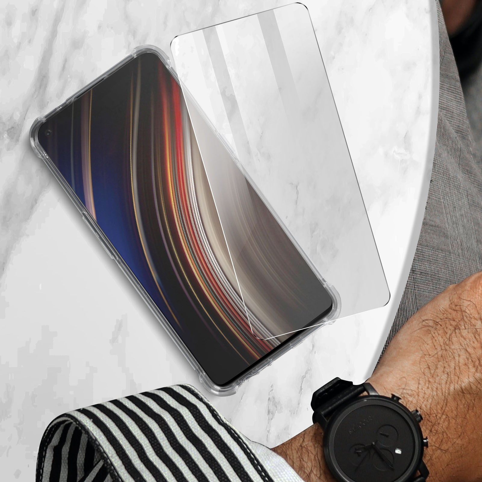 Ein Telefon mit Displayschutz und eine Uhr auf einem Tisch. Das Telefon hat ein farbenfrohes Display. Schwarze Uhr.