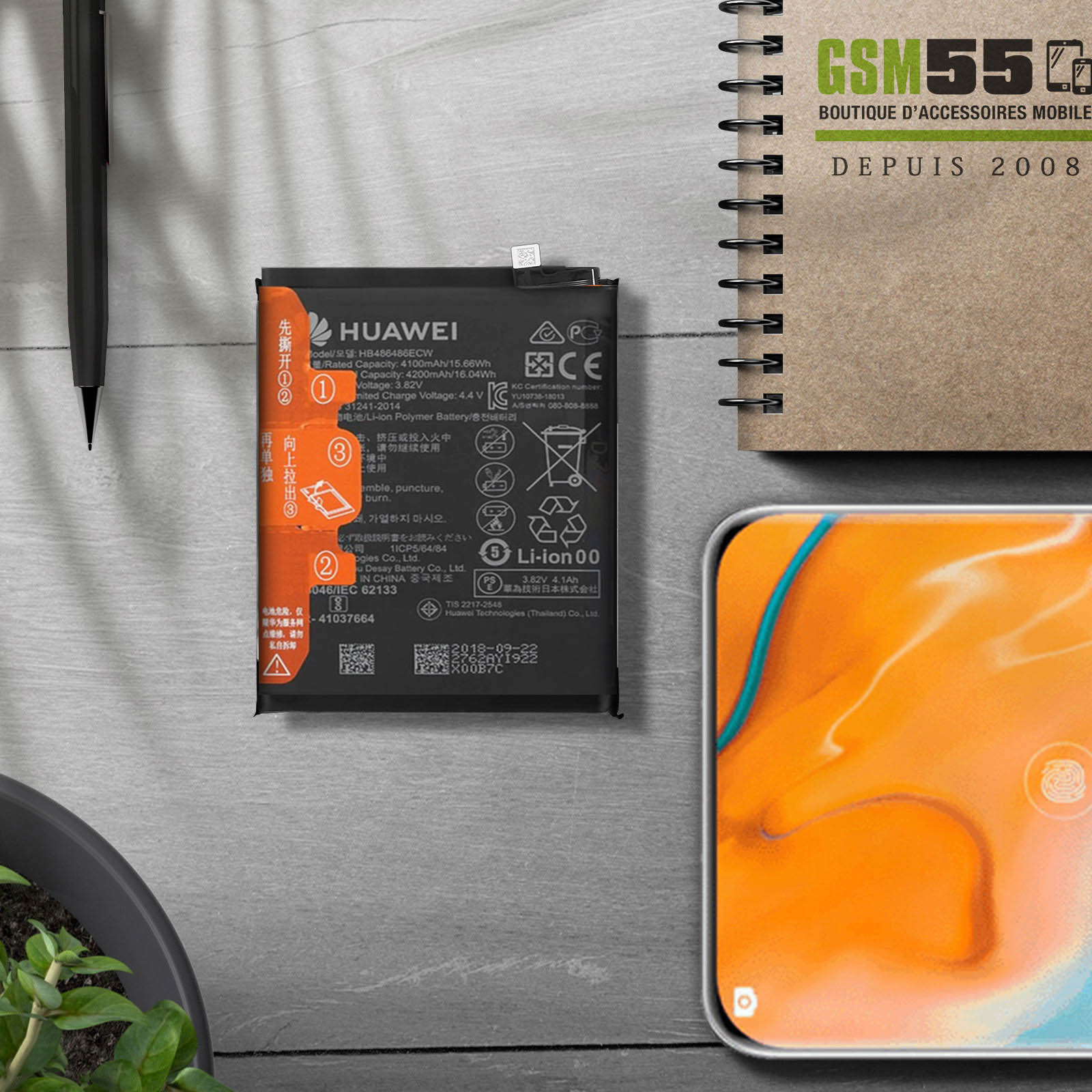 Batteria e telefono su una scrivania. La batteria ha testo e simboli. Scrivania con taccuino, penna e pianta.