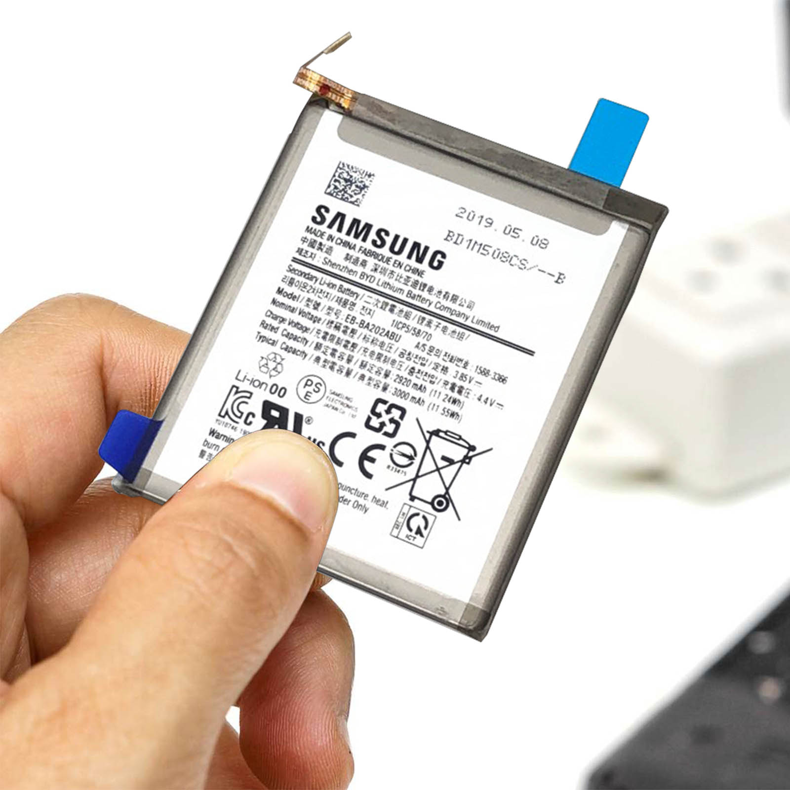 Main tenant une batterie Samsung avec des étiquettes, sur fond blanc et gris.