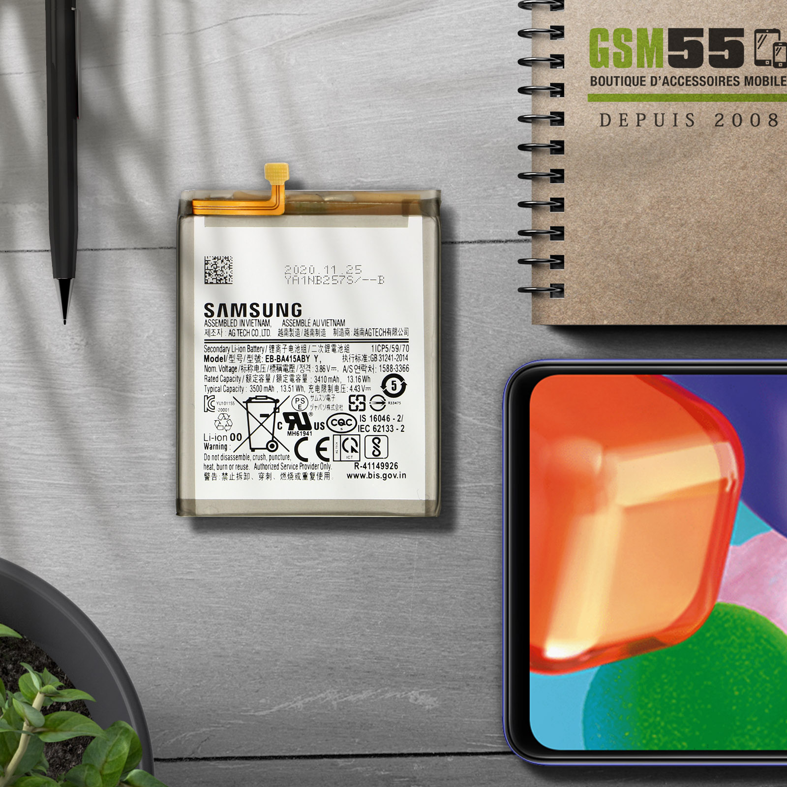 Une batterie de téléphone avec le logo SAMSUNG et une étiquette. La batterie est posée sur une table avec une plante.