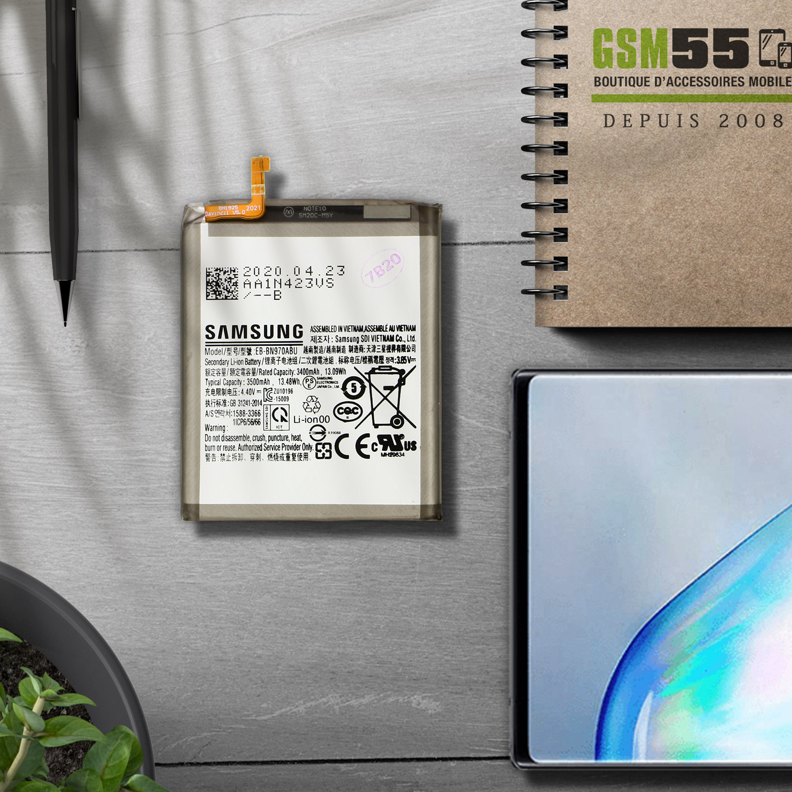 Ein Samsung-Akku mit Etiketten. Daneben befinden sich ein Telefon, ein Stift und eine Pflanze auf einem Holztisch.