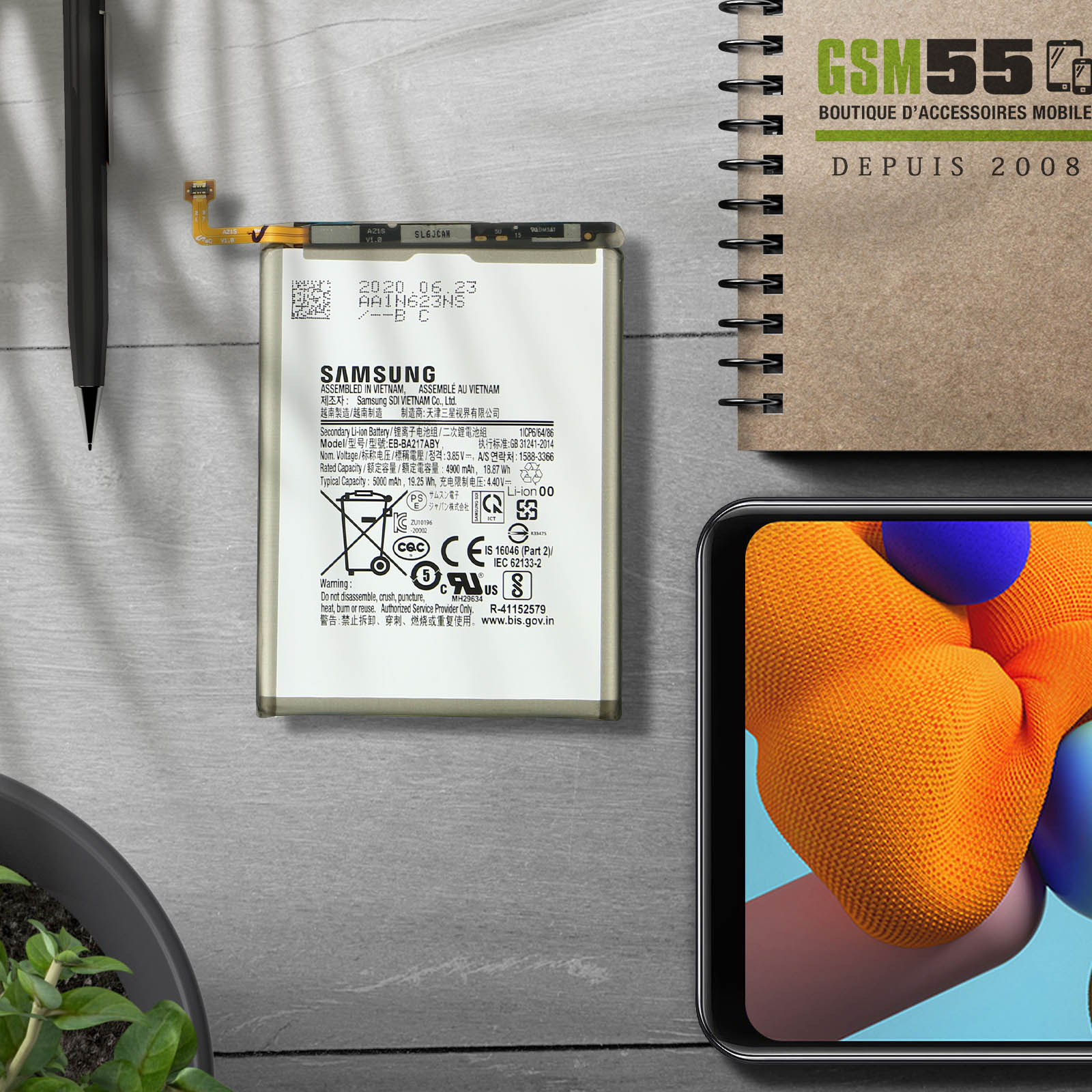 Batterie avec étiquettes et marquages de sécurité. Téléphone et accessoires sur un bureau. Le bureau a une plante et un carnet.