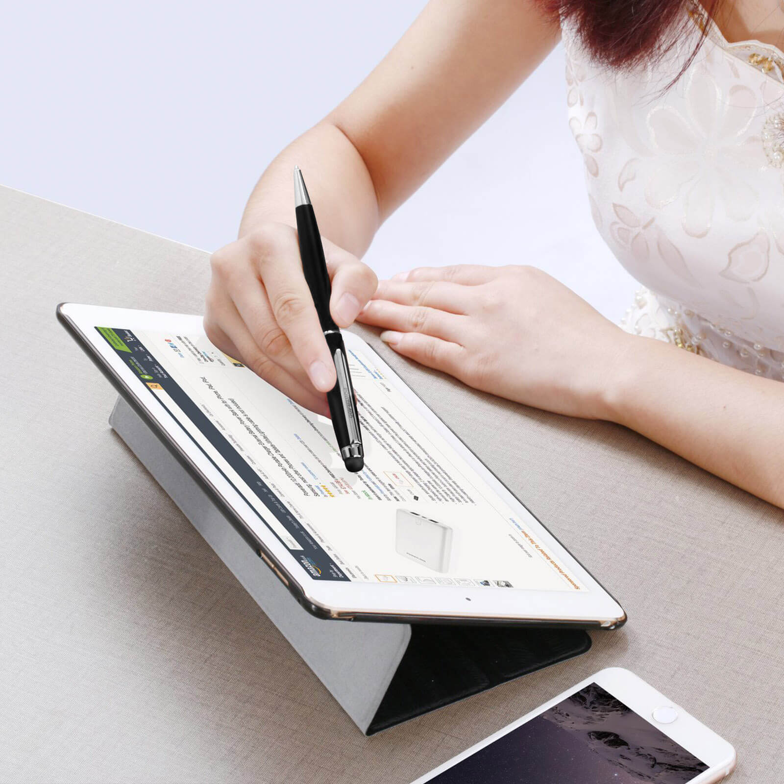 Una mano usa una penna nera su un tablet. Sotto c'è un telefono. La donna indossa un vestito bianco.