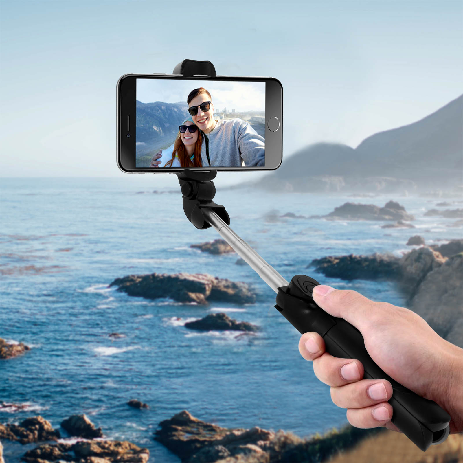 Mano che tiene un bastone per selfie nero con telefono che mostra una coppia. Sfondo oceano e montagne.