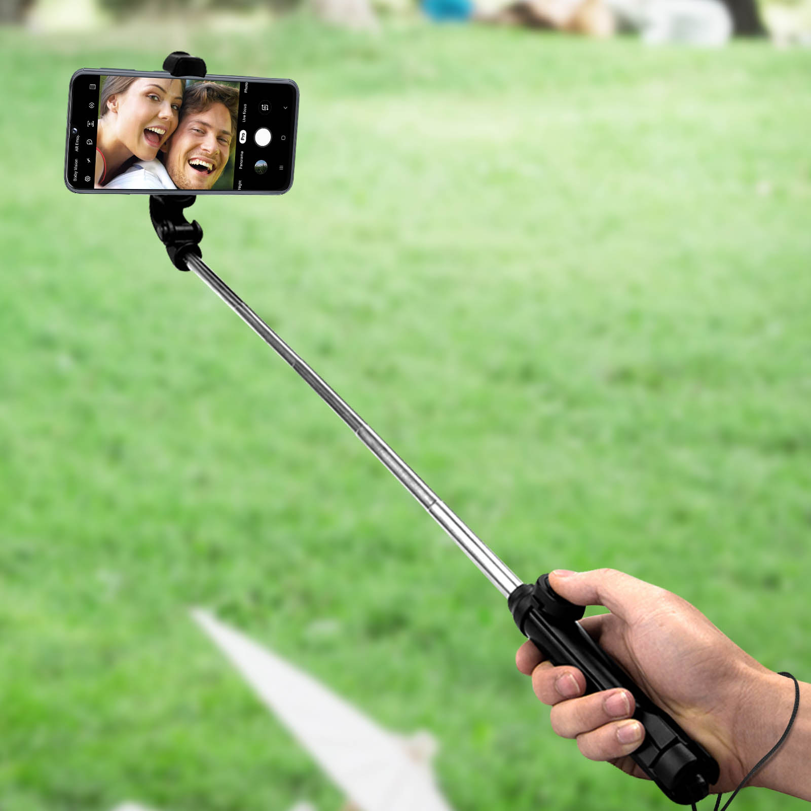 Hand hält Selfie-Stick mit Telefon, das ein Paar im grünen Gras zeigt.