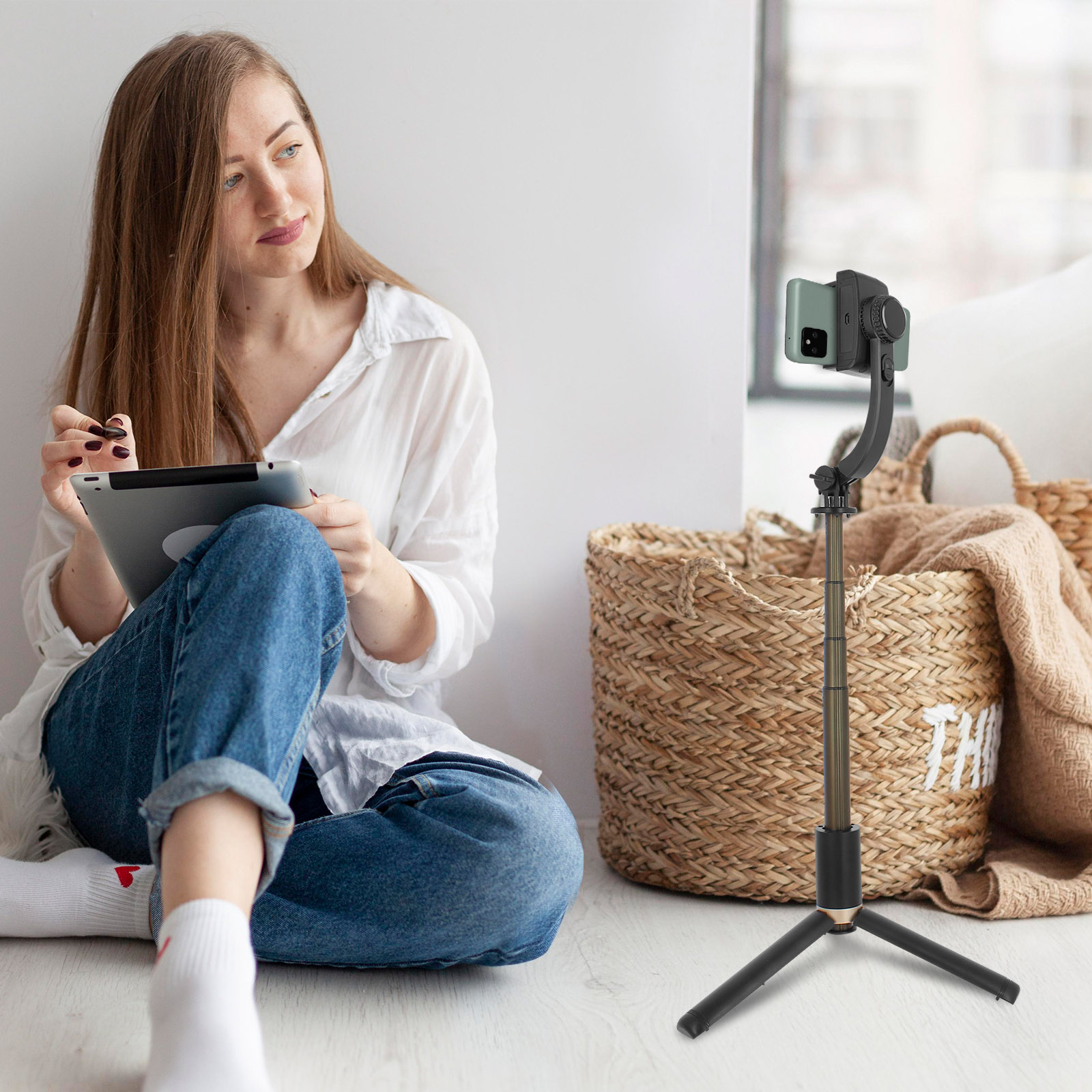 Mujer con tableta y un palo selfie con trípode con teléfono adjunto. Cesta y manta en el fondo.