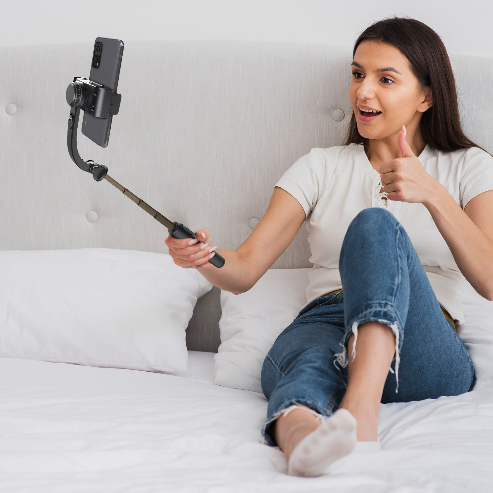 Mujer en cama con un palo selfie, pulgar arriba. Teléfono en el marco.