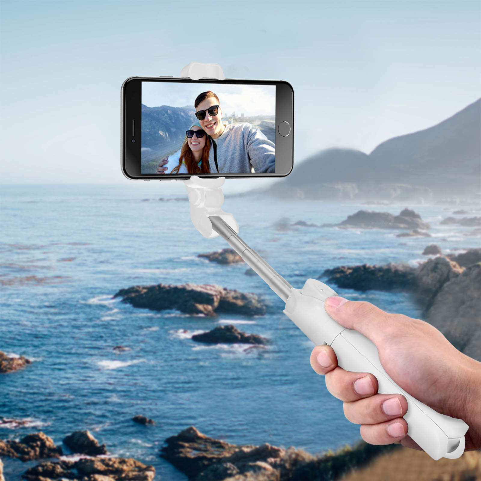 Mano sosteniendo un palo selfie blanco con teléfono que muestra una pareja. Fondo de océano y montañas.