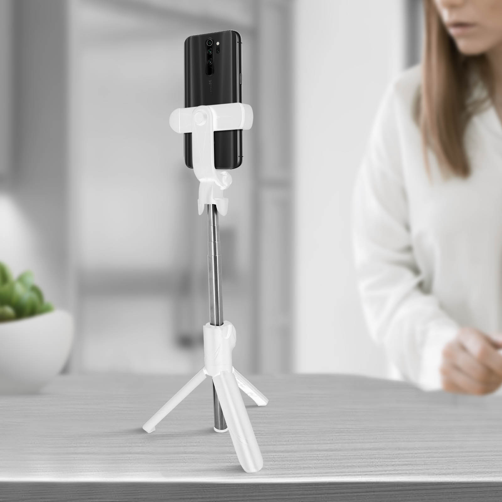 Un palo selfie blanco con un teléfono, sobre una mesa, con un fondo borroso de una cocina.