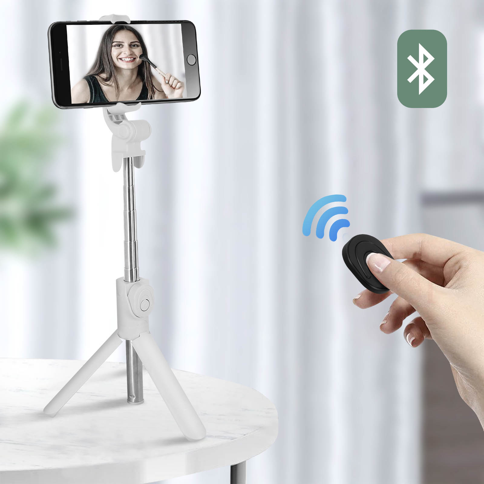 Teléfono en trípode blanco con mando Bluetooth. Mano sosteniendo el mando con señales azules.