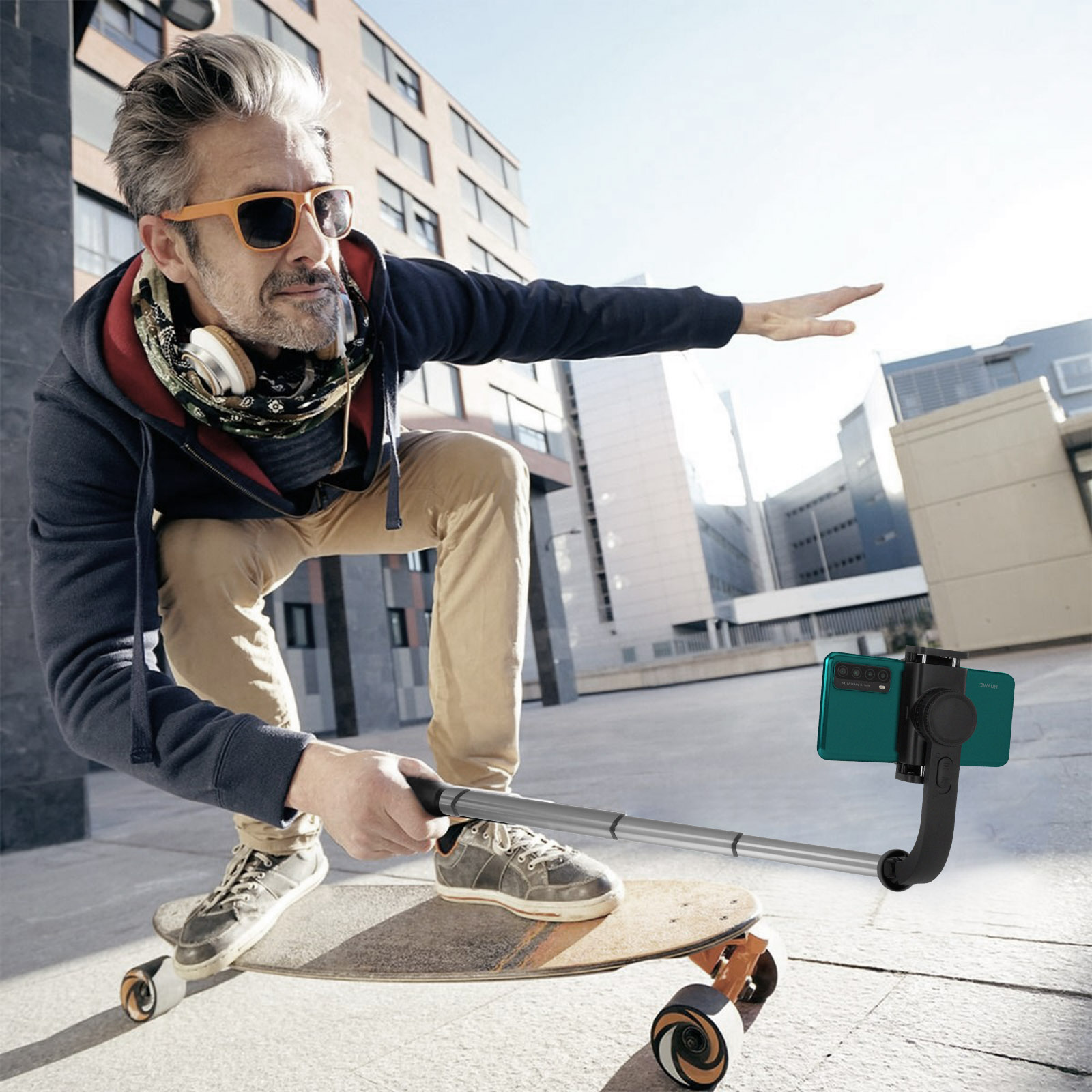 Un uomo su uno skateboard che usa un bastone per selfie con un telefono, all'aperto alla luce del sole.