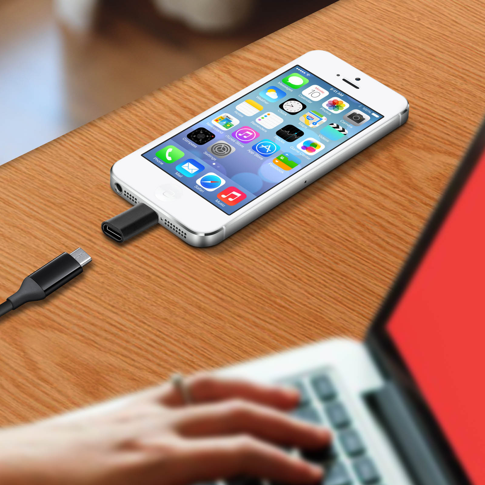 iPhone blanc connecté à un adaptateur noir sur table en bois, à côté d'un ordinateur portable. Main floue.
