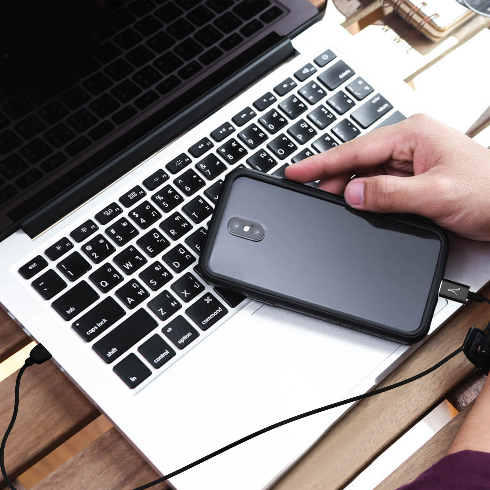 Una persona tiene in mano un telefono nero collegato a un laptop con un cavo nero. Il laptop è su un tavolo di legno.