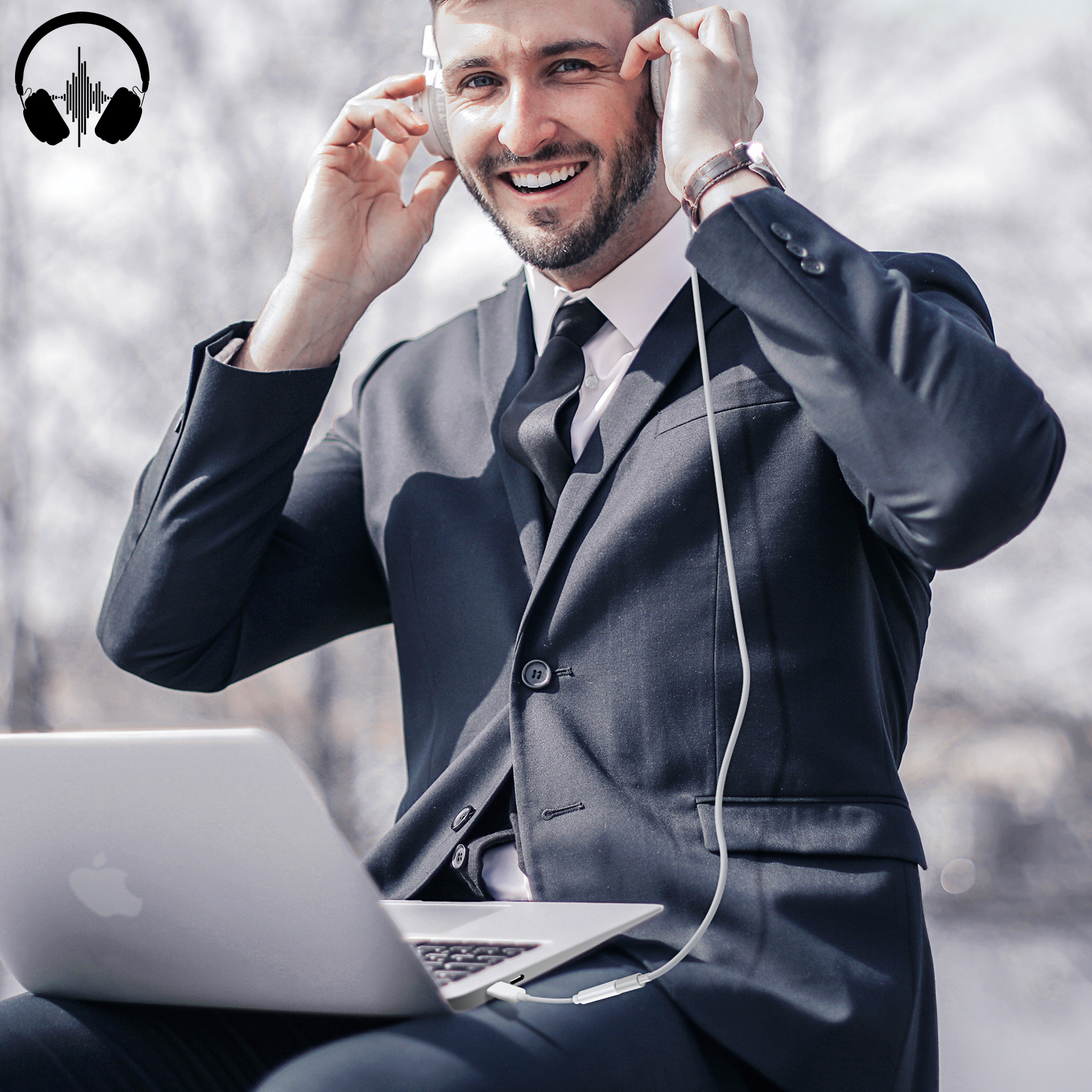 Hombre sonriente con auriculares, traje. Sostiene los auriculares mientras está sentado con un portátil.