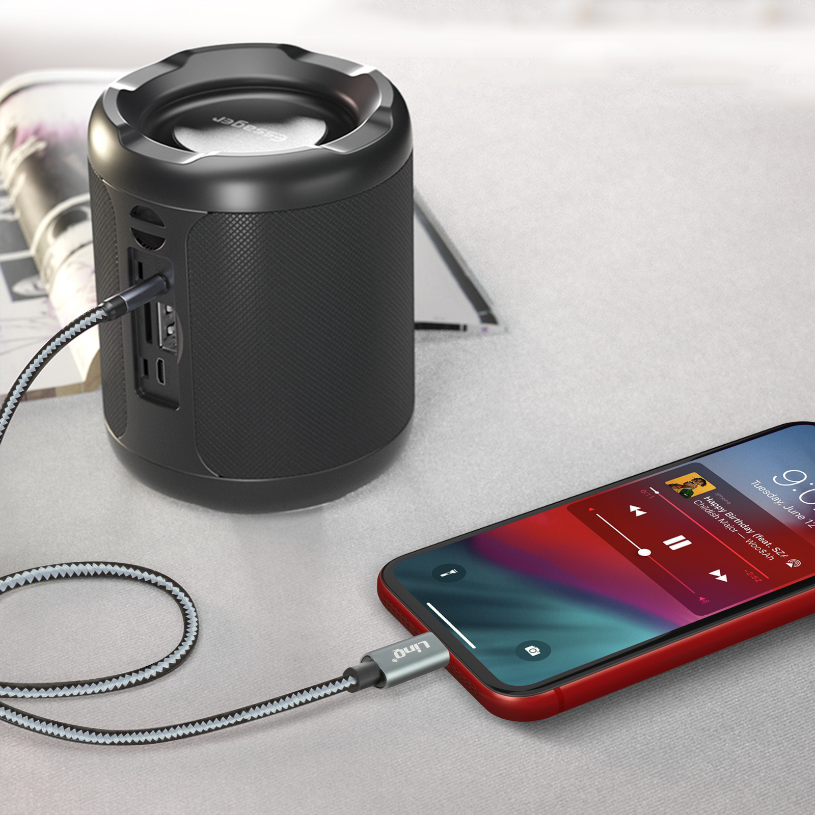 Altavoz negro junto a un teléfono con una funda roja. Un cable de carga está conectado. Superficie blanca.