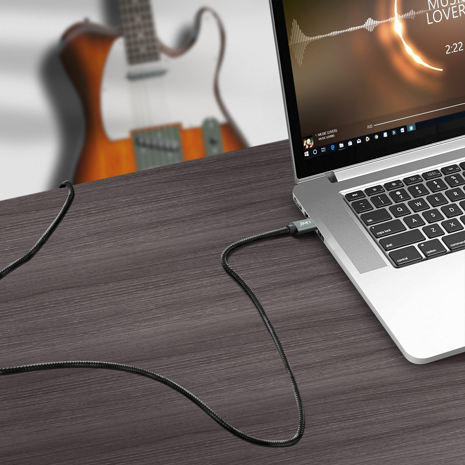 Una computadora portátil con un cable conectado, reproduciendo música. Una guitarra está en el fondo.