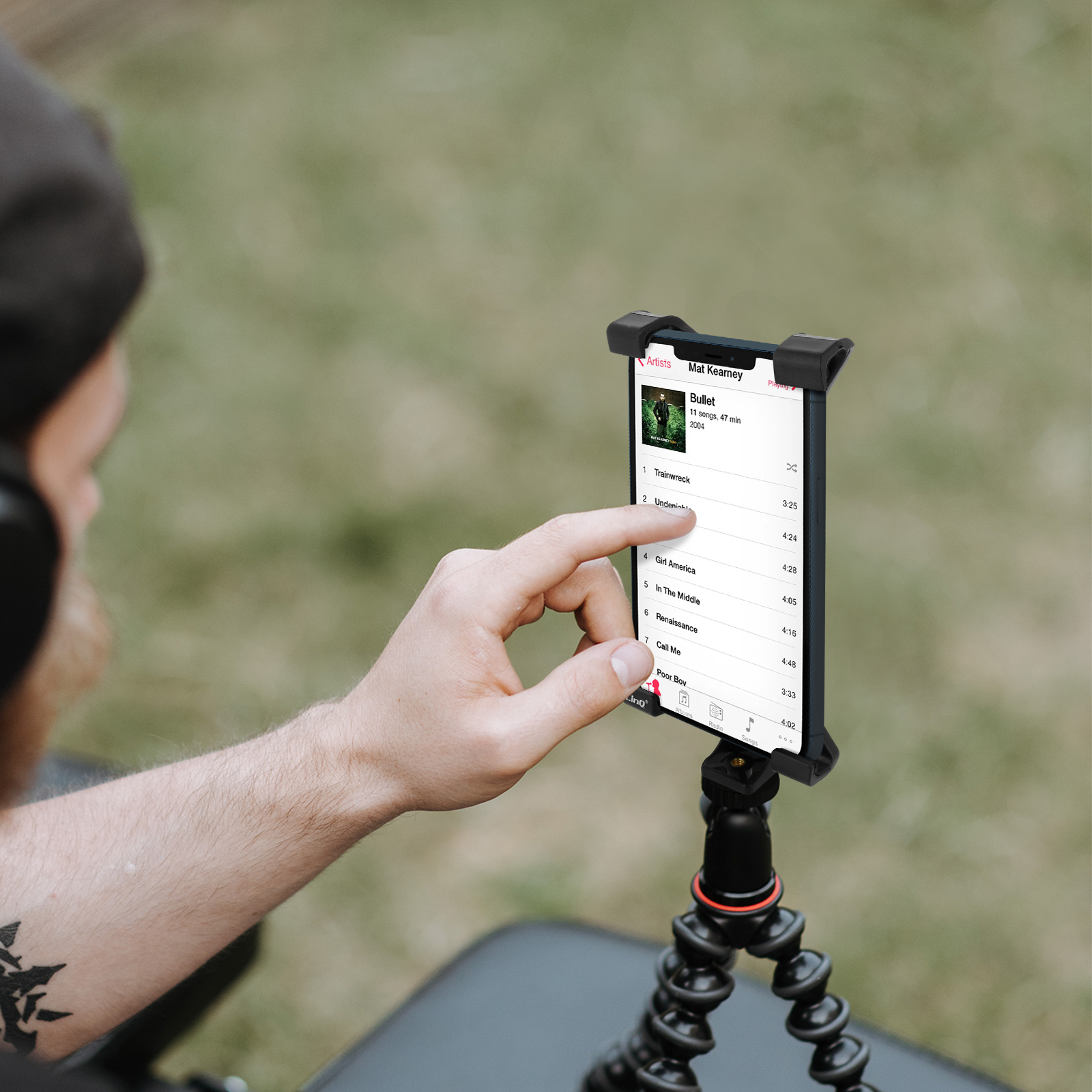 Persona che usa un tablet montato su un treppiede. Mostra una lista di canzoni. Sfondo di erba verde.