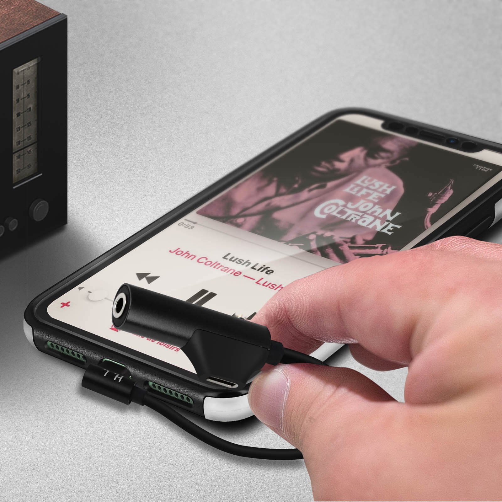 Mano insertando un adaptador negro en un teléfono. El teléfono está reproduciendo música. Fondo gris.