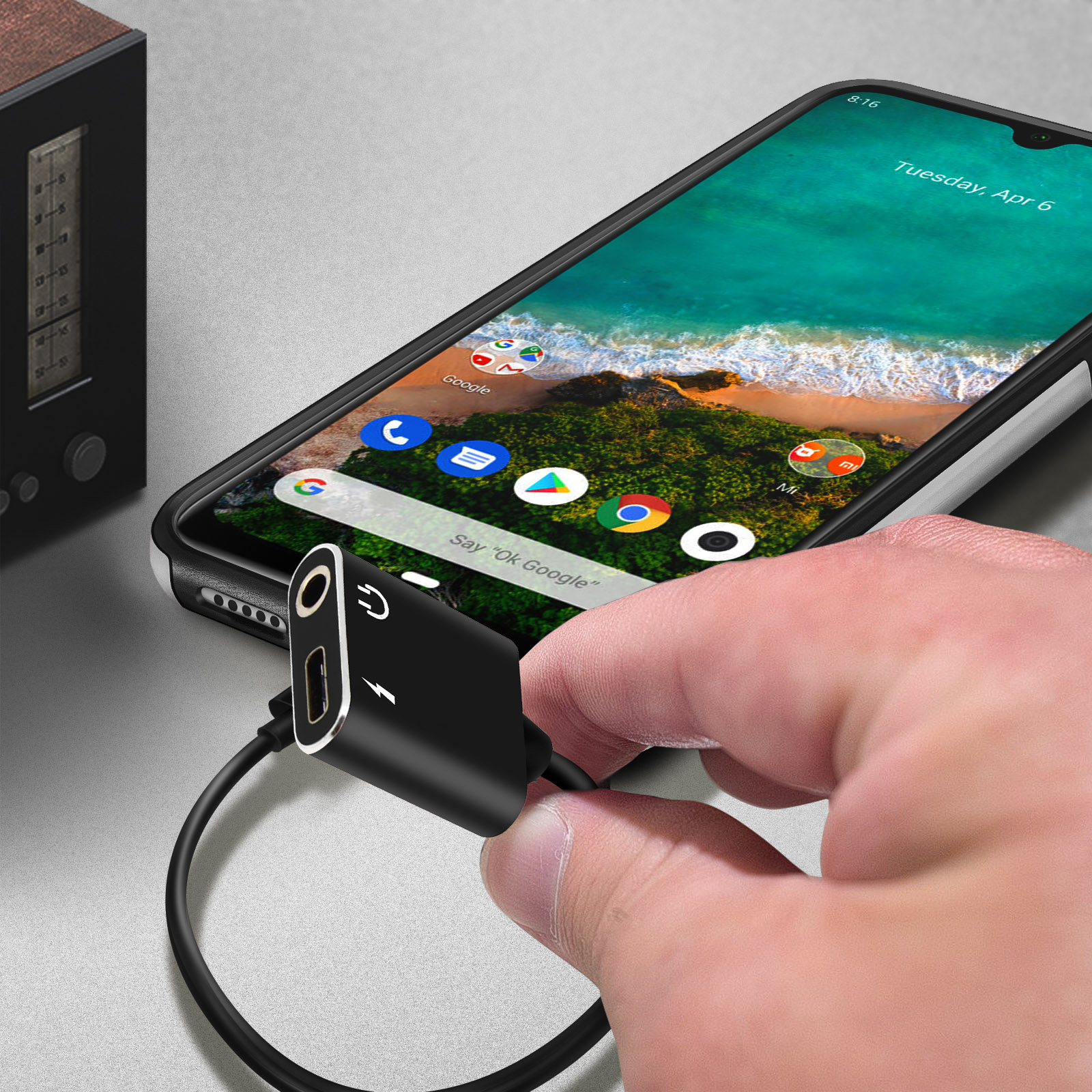 Un adaptador negro se sostiene con la mano, conectado a un teléfono y un cable. Una radio está en el fondo.
