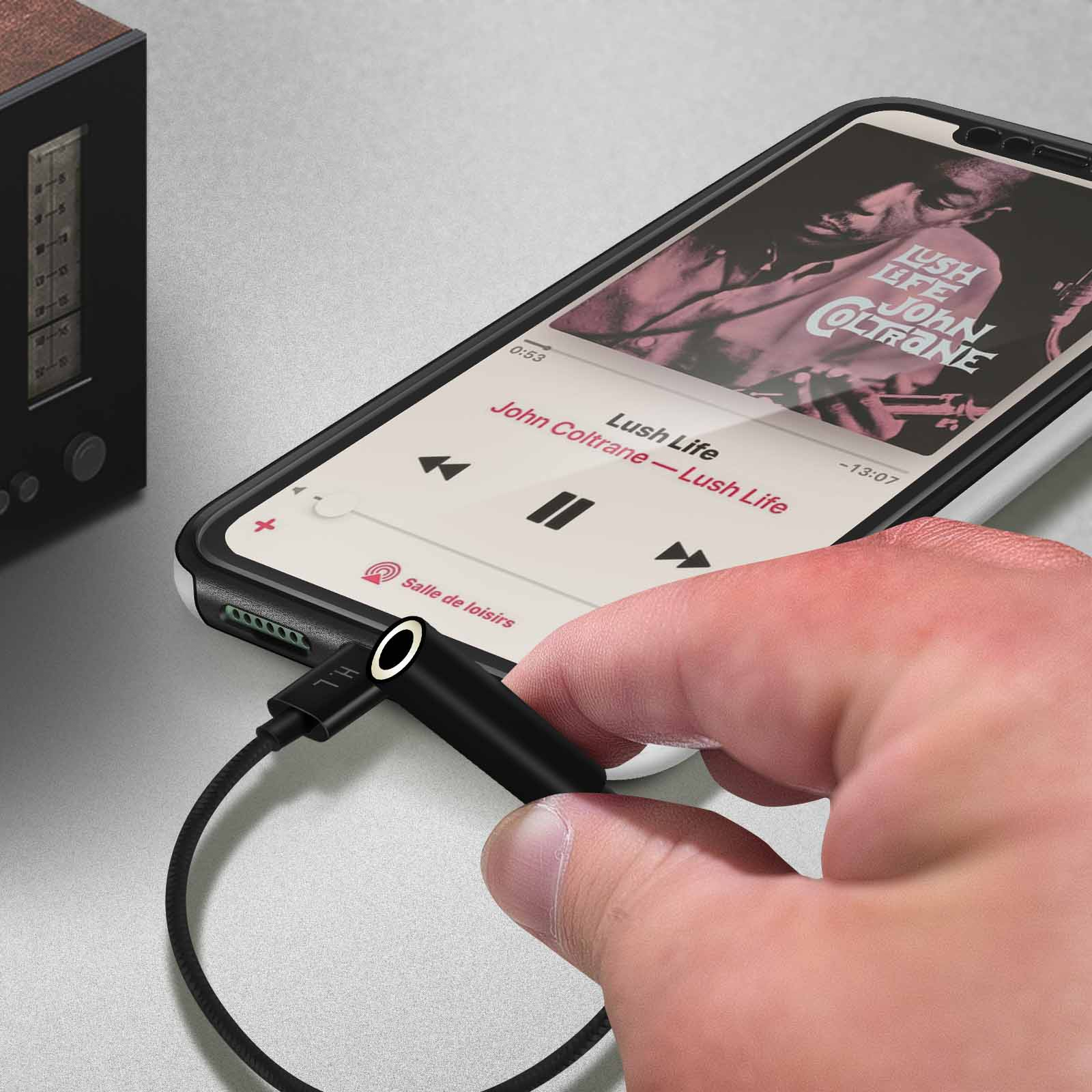 Una mano enchufando un adaptador de audio negro a un teléfono reproduciendo música, con una radio al fondo.