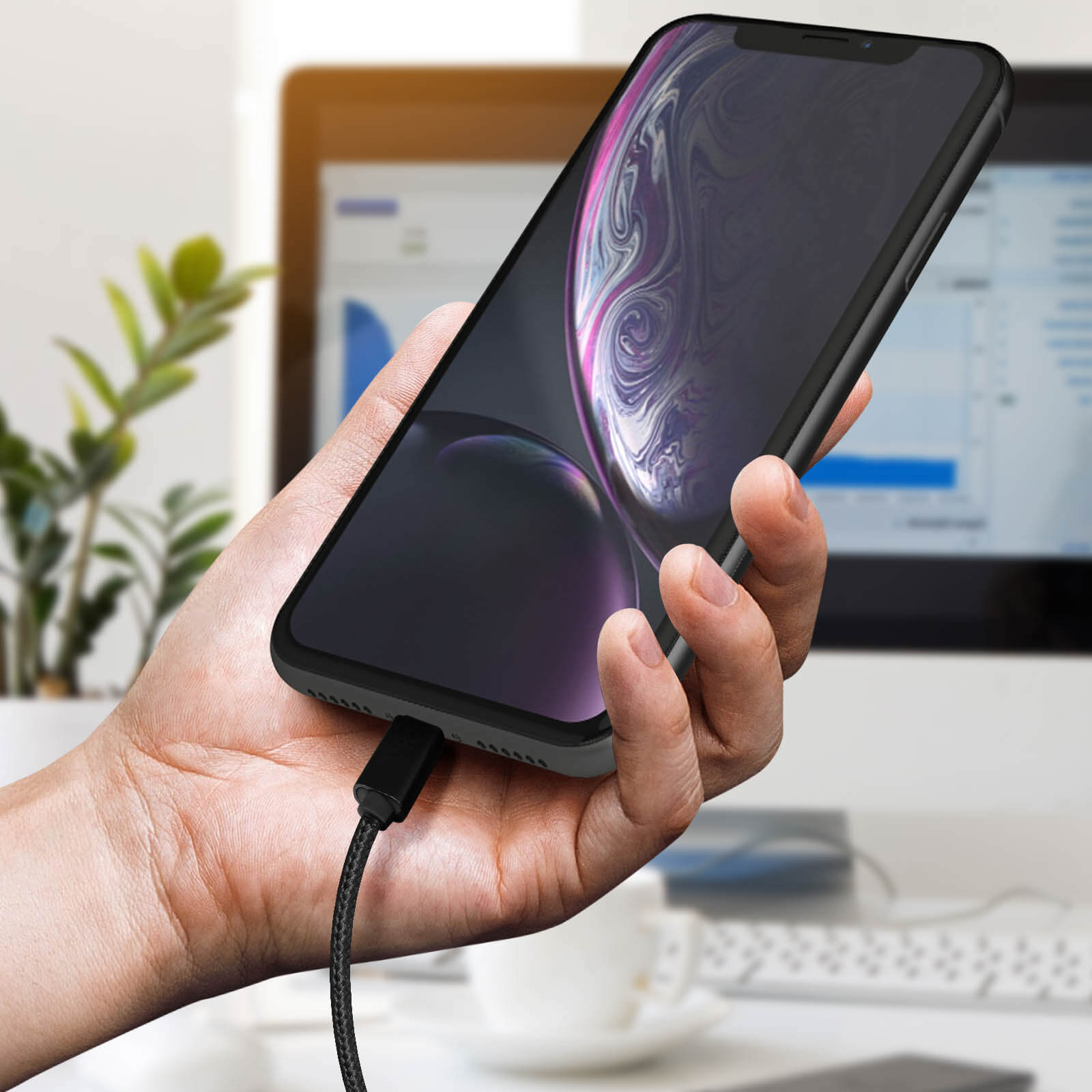 Una mano che tiene un telefono con un cavo collegato, un monitor del computer è sullo sfondo.